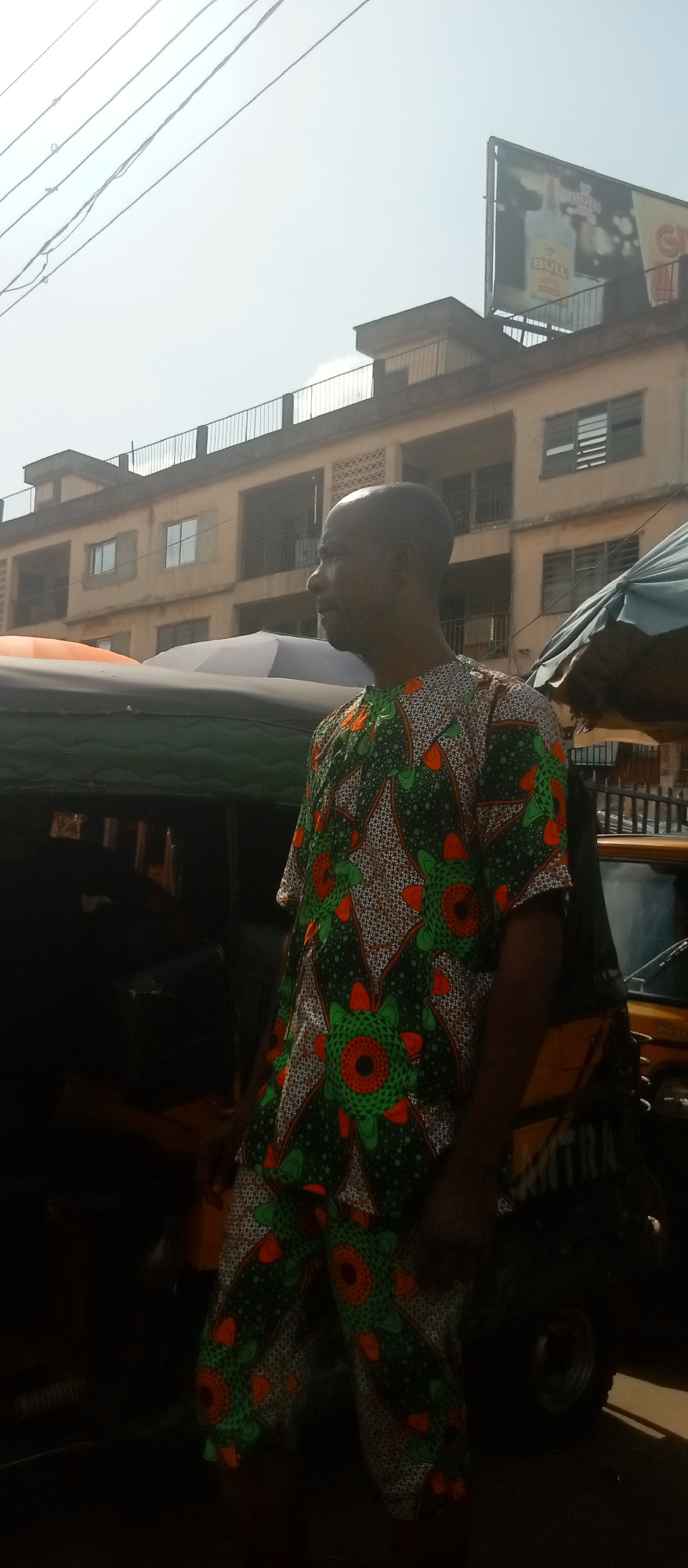 The image presents an outdoor scene in Nkpor, Nigeria. A man is prominently featured, wearing a short-sleeved, patterned shirt and matching trousers. He is situated near a motorized tricycle taxi, commonly known as a "Keke Napep," partially obscured behind him. Other parked vehicles are visible in the background. The backdrop includes a multi-story building and a billboard advertising a product called "BULL." Overhead, power lines crisscross the sky. The lighting suggests a daytime setting under a bright sun.