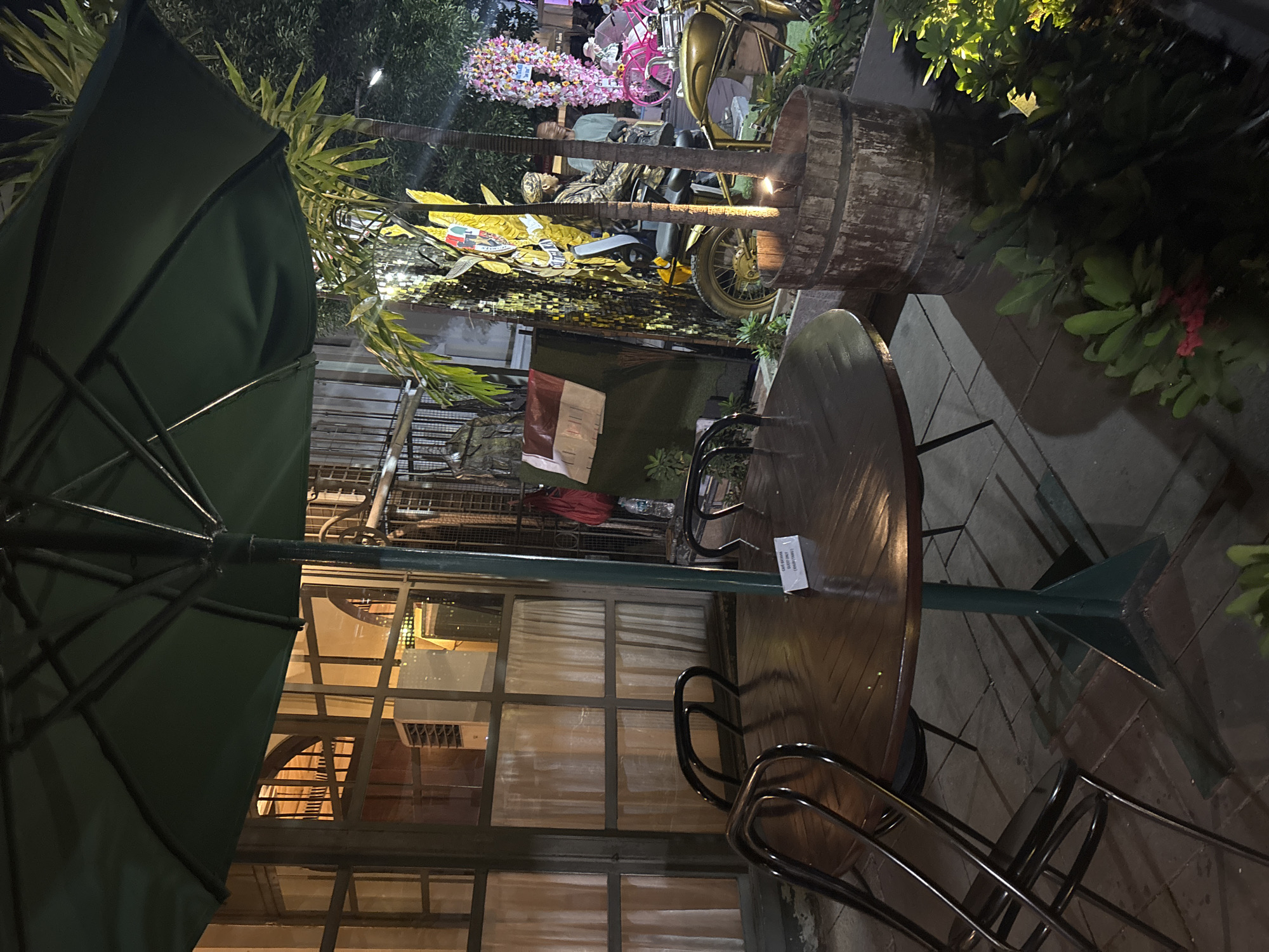 An outdoor seating area is depicted at night, possibly at a cafe or restaurant. In the foreground, a round wooden table is set with chairs. A sign on the table reads "CAFE BATHARA" with additional text below that is difficult to read. To the right of the table, a large wooden barrel planter contains two palm trees. Behind the planter, a motorcycle is visible, with a mannequin dressed in military attire seated on it. Further to the right, a display of pink flowers and a pink bicycle are present. The background includes a building with large windows and interior lighting, as well as other foliage and decorative elements. The overall scene suggests a lively atmosphere, possibly a place for outdoor dining or events.