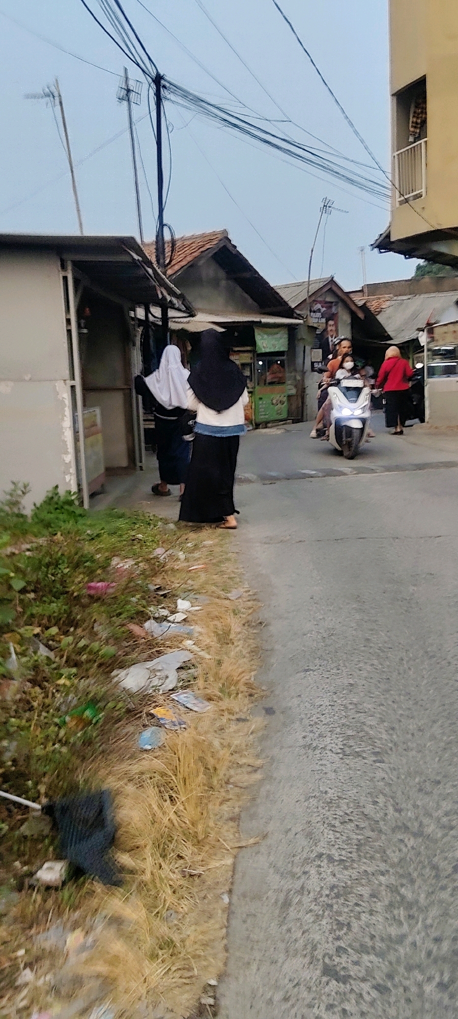 The image depicts a street scene in Mekarmukti, Indonesia, likely during the late afternoon or early evening due to the soft lighting. In the foreground, a grassy verge is littered with various pieces of trash, including plastic wrappers and what appears to be fabric. The verge transitions to a paved street.
On the street, two women wearing hijabs are walking away from the camera, one in a white hijab and the other in a black hijab. To their right, a motorcycle is being ridden by two individuals, a driver and a passenger, both wearing face masks. The driver is wearing an orange shirt, and the passenger is wearing a red top. Further down the street, another person in a red top and black pants is walking away from the camera.
The background features several small buildings with tiled roofs, some appearing to be storefronts. A complex network of electrical wires crisscrosses overhead, supported by poles. On the right side of the image, a section of a light-colored building with a balcony and laundry hanging on it is visible. The overall environment suggests a residential or commercial area with typical urban infrastructure.