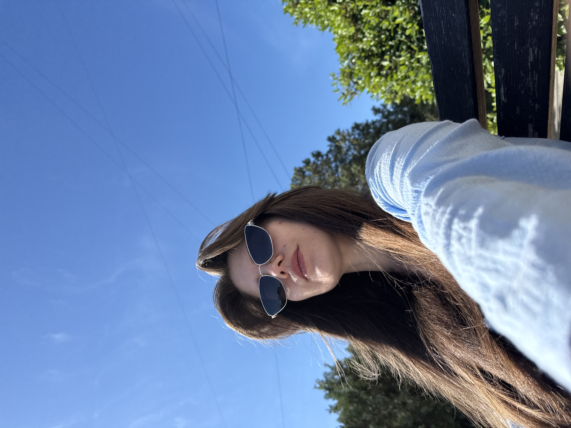 A medium shot captures a young woman from the chest up, seated outdoors on a park bench. She is facing slightly away from the camera, with her head tilted upwards towards a bright blue sky. Her long, brown hair is partially obscuring her face, with strands caught in a breeze. She is wearing a light blue long-sleeved shirt and silver-framed sunglasses. The background consists of green foliage from trees and the clear sky, with faint lines that could be power lines or contrails. The bench she is sitting on appears to be dark wood. The lighting suggests it is a sunny day. The location is identified as Unknown, Unknown.