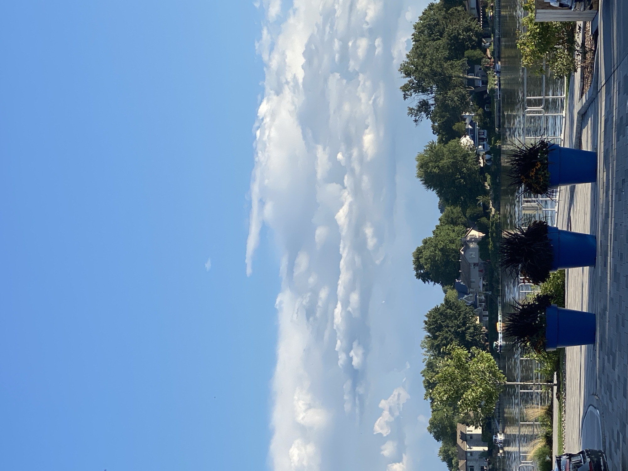 The image depicts a waterside scene with three large blue planters in the foreground, each containing dark-leafed plants and some yellow flowers. Beyond the planters, a body of water, likely Lake Saint-Louis, is visible, with boats and houses on the opposite shore. The houses are nestled amongst lush green trees. The sky above is a vibrant blue, with scattered white clouds. The perspective suggests a paved walkway or promenade in the foreground, with a modern railing system bordering the water. The overall impression is one of a serene, well-maintained urban park or waterfront area in Sainte-Anne-de-Bellevue, Canada.