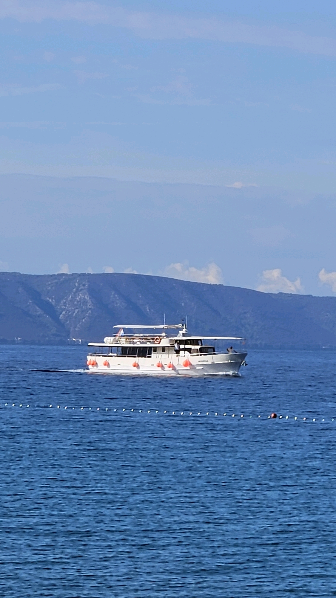 A white passenger ferry with multiple decks moves across a deep blue sea under a pale blue sky. The ferry has several windows and railings, and bright orange buoys are visible along its side. Two people are visible on the top deck of the ferry, one standing and one sitting. In the background, a dark blue mountain range with visible slopes rises from the sea, with a few scattered white clouds in the sky above it. A line of small white buoys with one red buoy in the distance marks a boundary in the water closer to the foreground. The scene appears to be in Općina Tučepi, Croatia, given the coastal mountain range and blue waters characteristic of the region.