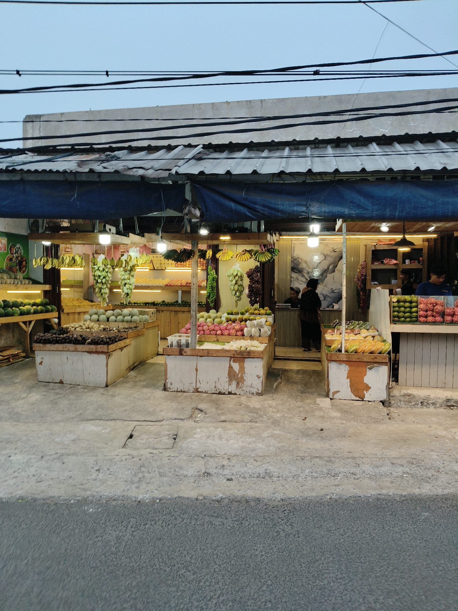 A fruit stall is depicted on a street in South Tangerang, Indonesia. The stall is covered by a blue tarp and features a variety of fruits displayed on wooden stands. Fruits such as watermelons, melons, mangoes, dragon fruits, bananas, and durians are visible. The stall is illuminated by several lights. Two individuals are working behind the counter, and a customer is present. The street in front of the stall is paved with concrete and asphalt. The sky above is overcast.