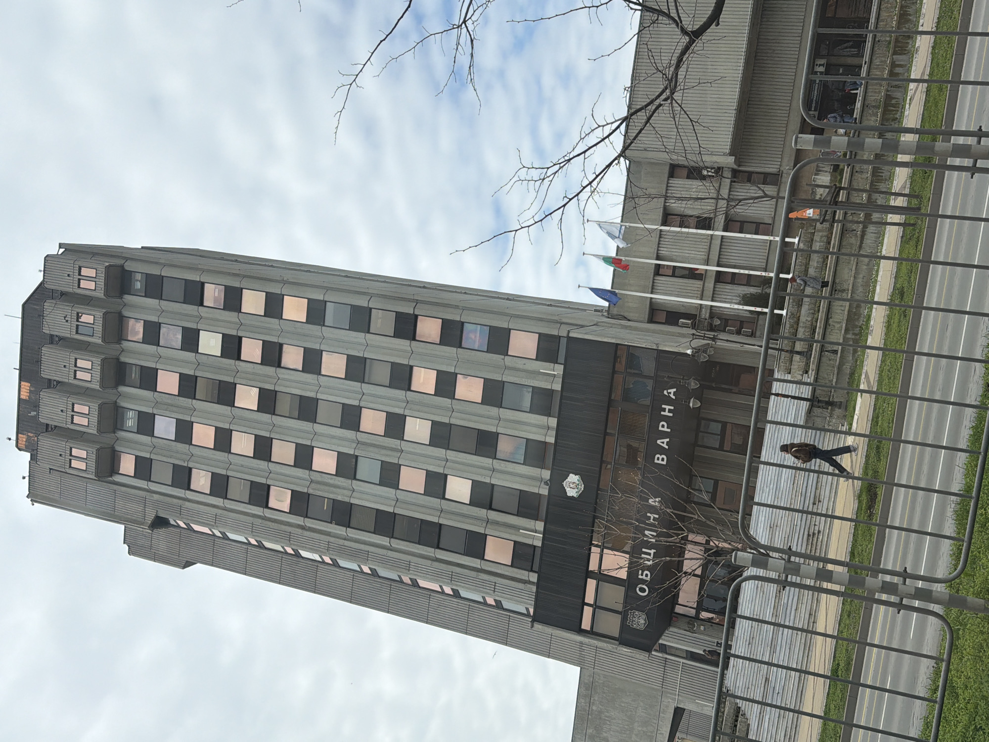 The image shows the Municipality of Varna building in Varna, Bulgaria. It's a tall, modernist structure with rows of windows. The building's facade includes the inscription "ОБЩИНА ВАРНА" (Municipality of Varna) in Cyrillic. Several flags are flying near the building. A metal fence runs along the front of the building, separating it from the street. A person is walking on the sidewalk behind the fence. The sky is overcast with a mix of clouds. Bare tree branches are visible in the foreground and around the building.
