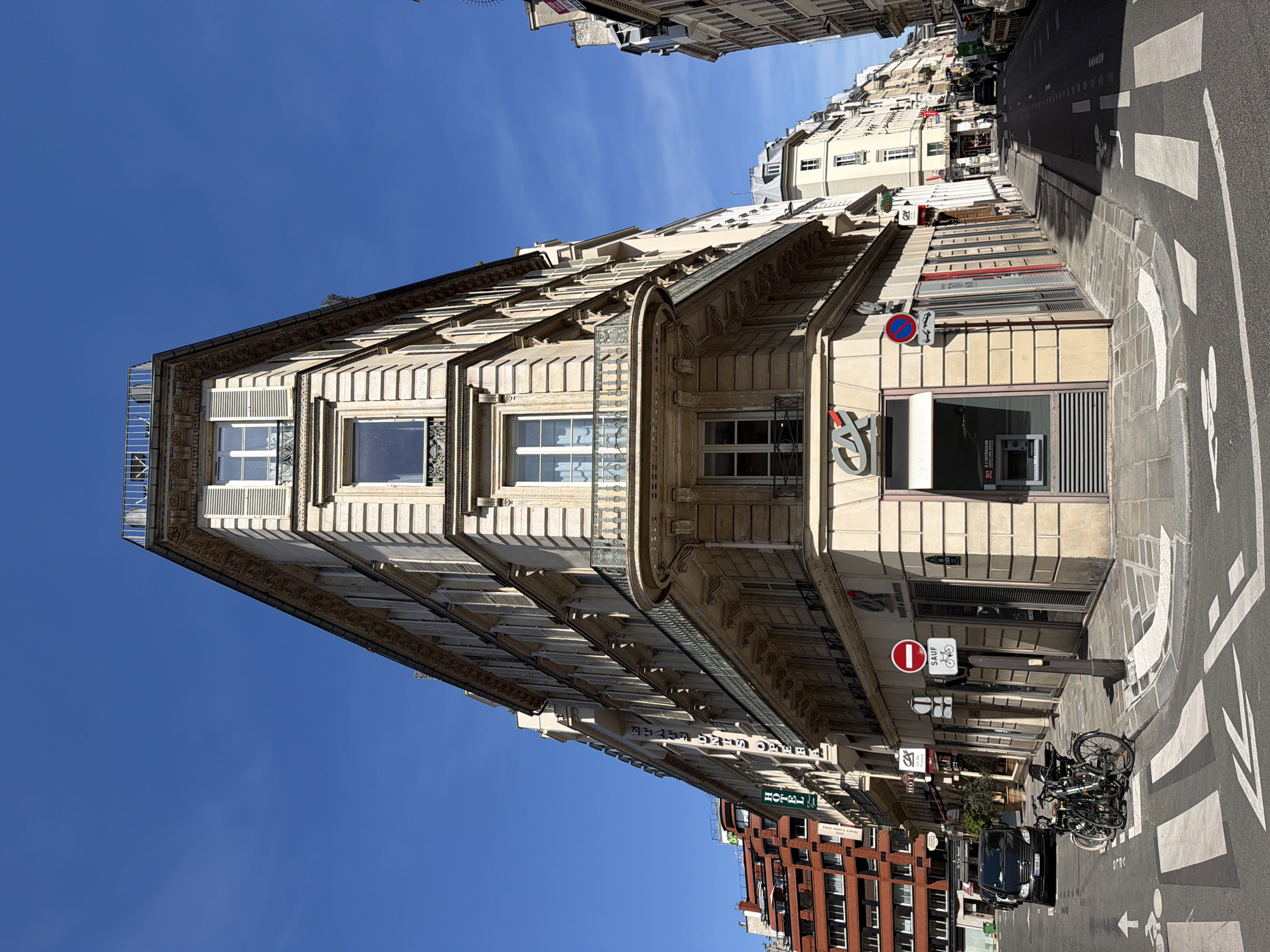 The image depicts a wedge-shaped building in Paris, France, known as the "flatiron building" or "L'Equerre." The building has a distinctive triangular facade, typical of structures built on converging streets. The architecture features classical details such as window frames, balconies, and ornate cornices. The building's street level appears to be a bank, indicated by the "Credit Agricole" logo and ATM. Street signs, a pedestrian crossing, and parked bicycles are visible, suggesting an urban setting. The sky is a clear blue. Other buildings line the street, indicating a dense, urban environment.