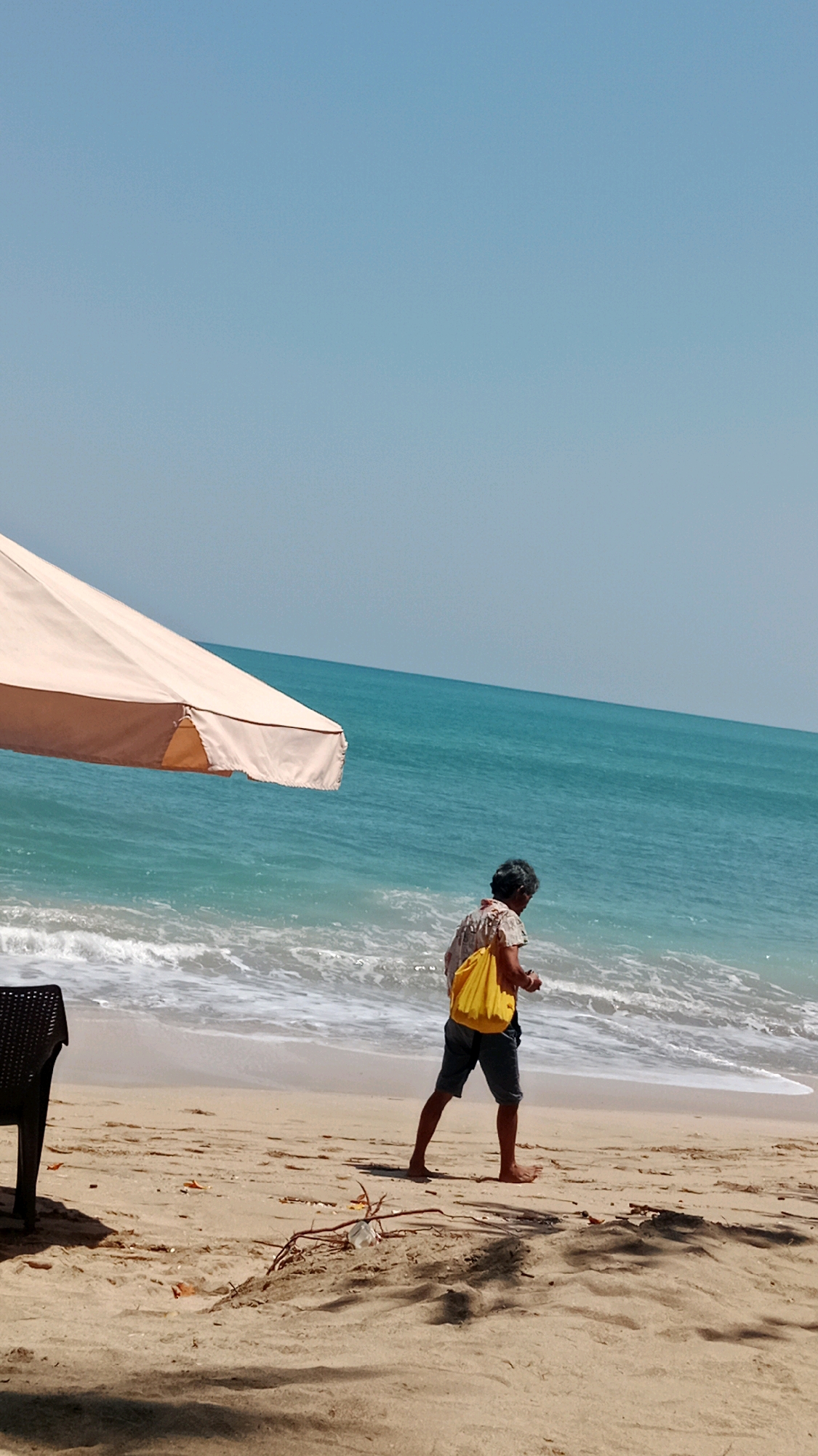 A person walks along a beach in Kuta, Indonesia. The person is wearing a patterned shirt, dark shorts, and is carrying a yellow bag over their right shoulder. They are walking towards the right of the frame with their back mostly to the viewer. The beach is sandy with some scattered debris. Waves are breaking on the shore. In the foreground, a portion of a beige beach umbrella and a dark chair are visible. The ocean is a vibrant turquoise color, and the sky is a clear, light blue.