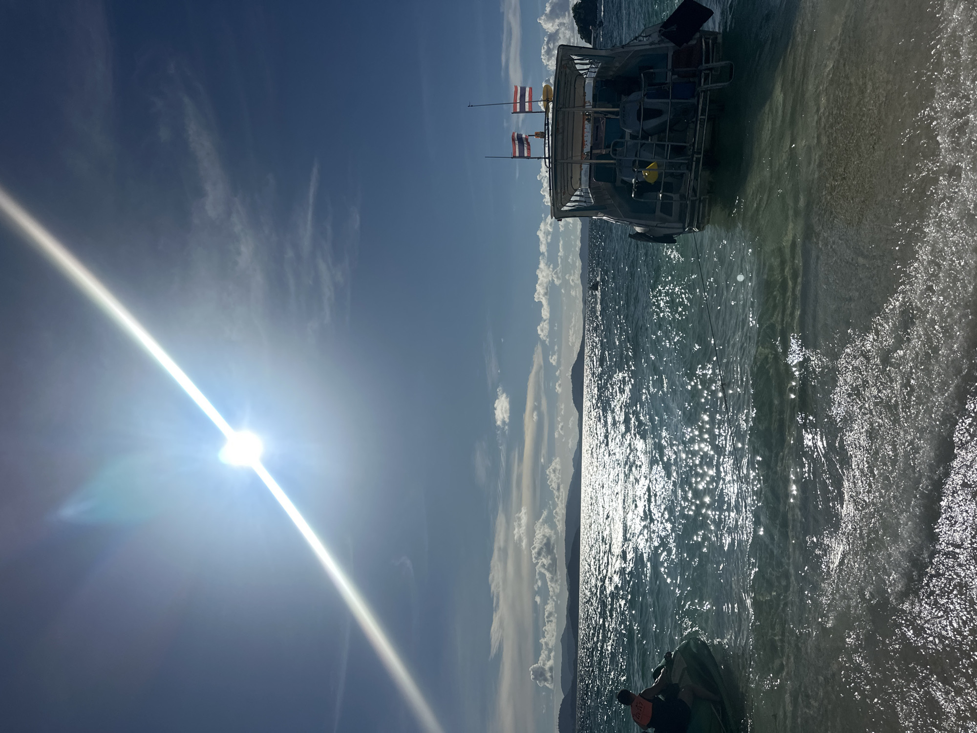 The image depicts a bright, sunny day at a tropical beach, likely in Phru Nai, Thailand. The sun is a prominent element, casting a strong glare across the scene and creating shimmering reflections on the water. The sky is a vibrant blue, dotted with fluffy white clouds.
In the foreground, the shallow, clear turquoise water gently laps at a sandy shore. A portion of a boat is visible, moored close to the beach. The boat appears to be a traditional Thai longtail boat, or a similar vessel, with an open deck and a canopy. Two Thai flags are prominently displayed on poles at the rear of the boat, suggesting its nationality.
Further out, the water extends to the horizon, where a silhouette of lush green hills or islands can be seen against the bright sky. The water itself is alive with the sparkle of sunlight, indicating a calm sea.
In the very bottom right corner of the image, a person is partially visible, seated on what appears to be a jet ski. They are wearing an orange life vest, suggesting preparation for water activities. The overall atmosphere is one of leisure and enjoyment of a beautiful day in a coastal setting. The time of day appears to be late morning or early afternoon, given the high position of the sun and the intensity of the light.