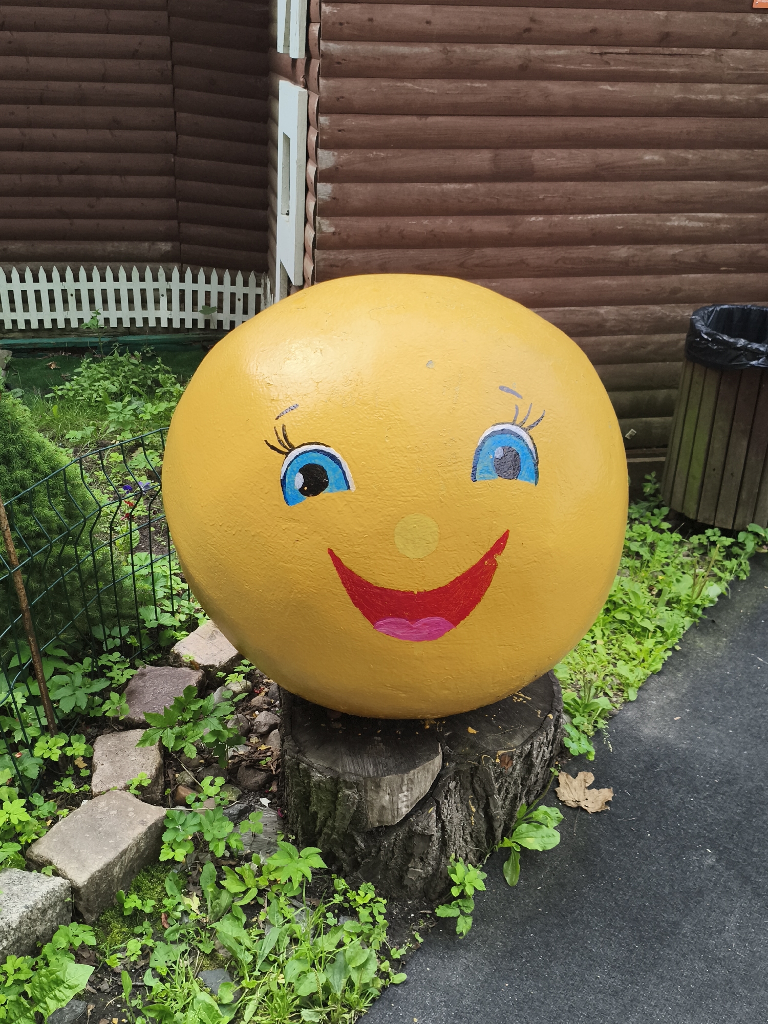The image shows a large, yellow, spherical object painted to resemble a smiley face. It is placed outdoors, resting on a tree stump in front of a brown wooden building with horizontal siding. A white picket fence is visible in the background. The ground around the object is a mix of asphalt and greenery, with some stone pavers and a metal fence. A black trash can with a liner is situated to the right of the smiley face. The lighting suggests it is daytime. No specific location in Kaliningrad, Russia, can be determined from the image itself.