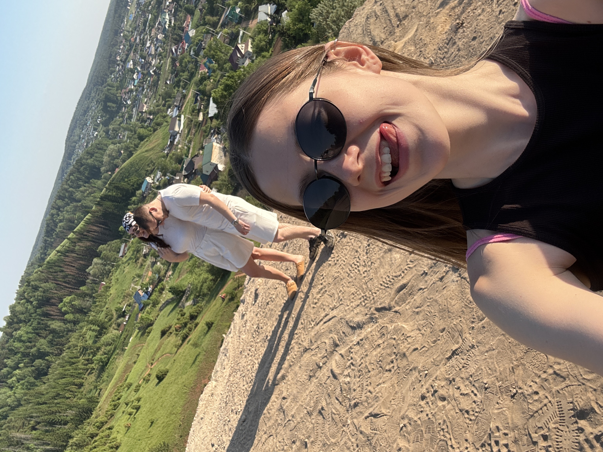 A selfie shows a young woman with sunglasses sticking her tongue out. She is wearing a black top with pink straps. Behind her, two people are standing closely together in white outfits. The location appears to be a raised sandy area with a view of the city of Ольховка, Russia, in the distance, showing rooftops and trees. The ground is marked with numerous footprints.
