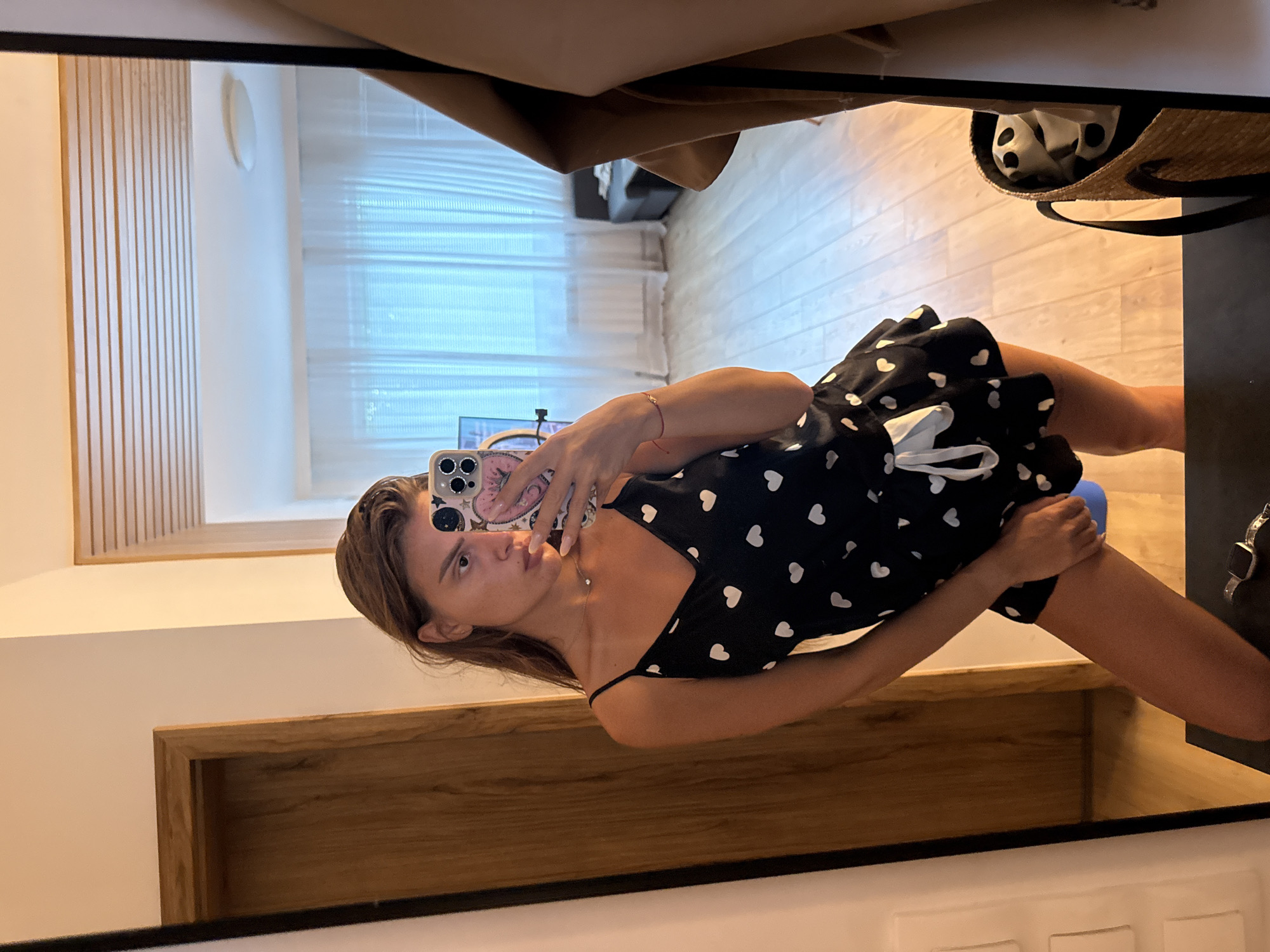 A young woman is taking a mirror selfie in what appears to be a modern apartment or hotel room. She is wearing a black sleeveless pajama set with white heart prints and a white ribbon tie at the waist. The pajama set has a V-neck and a ruffled hem. She is holding a smartphone in her right hand, which is decorated with colorful stickers. Her left hand is near her thigh, holding a blue object. She has long brown hair and is wearing a delicate necklace.
The mirror reflects the room, showing a wooden door on the left, a wall with horizontal wooden panels on the top, and a window with sheer white curtains in the background. Through the window, a blurred view of what appears to be a couch and possibly another room can be seen. The floor is made of light-colored wooden planks. A woven straw bag with black and white patterned fabric is visible in the lower right corner of the frame. A tan jacket or coat is hanging to the right of the mirror.
The environment suggests an indoor setting, likely a bedroom or living area. There are no identifiable landmarks or details that would specifically place this scene in Plovdiv, Bulgaria. The overall impression is one of casual domesticity or a vacation setting.