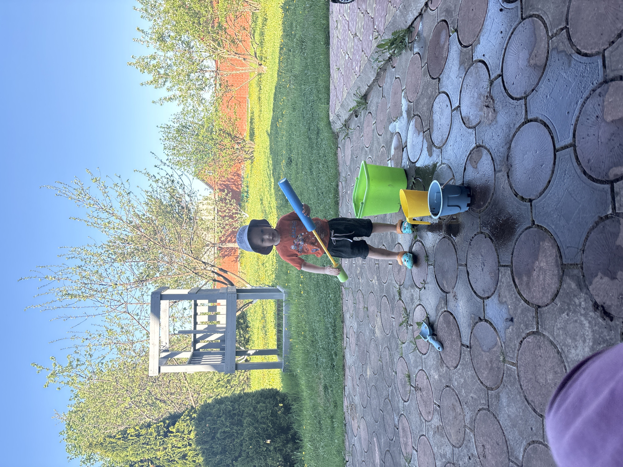 A young boy stands on a brick patio in Ферма, Russia. He wears a hat, an orange shirt, black shorts, and blue sandals. He holds a blue pool noodle and a yellow toy. Nearby are several colorful buckets and a small blue shovel. The patio is wet and surrounded by grass and trees. In the background, a wooden structure and a red brick building are visible. The sky is blue and clear.