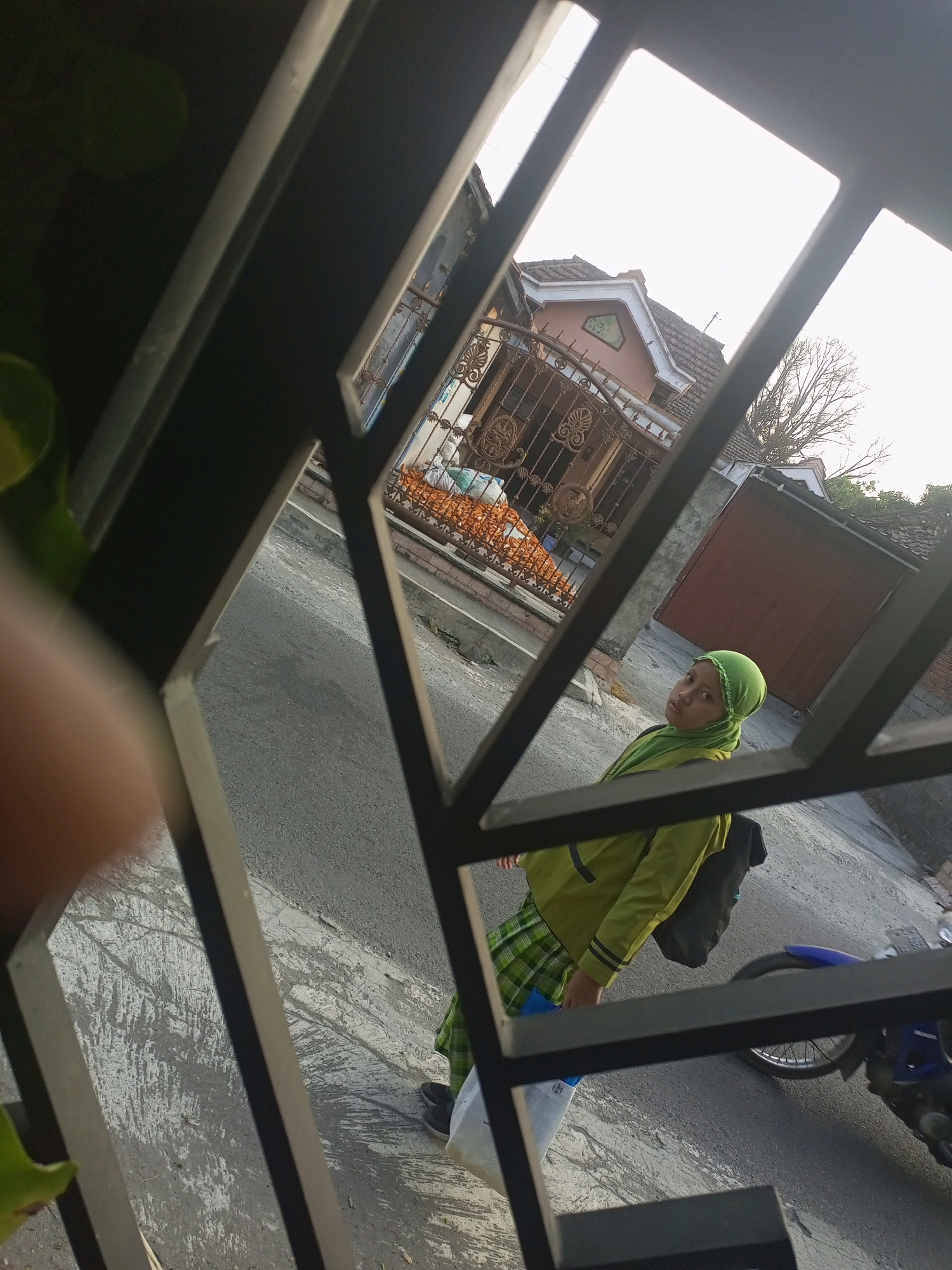 A young girl wearing a green hijab and a lime green jacket is standing on a paved street in front of a house in Ngasem, Indonesia. She is looking towards the camera. Her jacket has black stripes on the sleeves, and she is wearing a green and blue plaid skirt. She has a black backpack on her back and is holding a white plastic bag. A blue motorcycle is parked behind her. The house has a brown roof and a decorative metal fence. The sky is overcast.