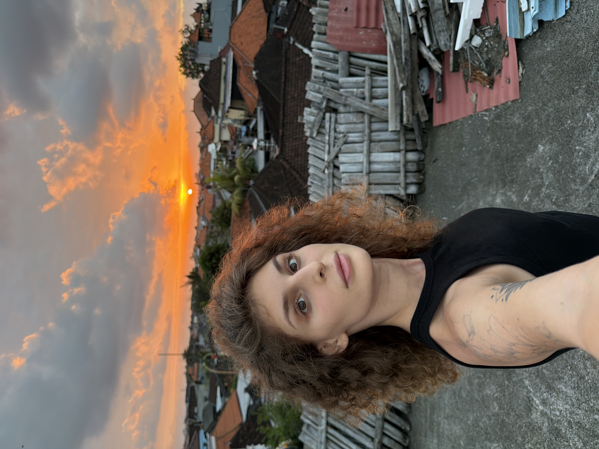 A woman with curly brown hair and a black tank top stands on a rooftop in North Kuta, Indonesia, during sunset. The sky is filled with dramatic orange and gray clouds as the sun dips below the horizon, casting a warm glow over the cityscape of rooftops. A wooden fence made of horizontal planks stretches across the middle ground, separating the foreground from the background. In the lower right corner of the image, a pile of discarded building materials, including wood planks and red corrugated metal, is visible. The woman is looking directly at the camera, her expression neutral.