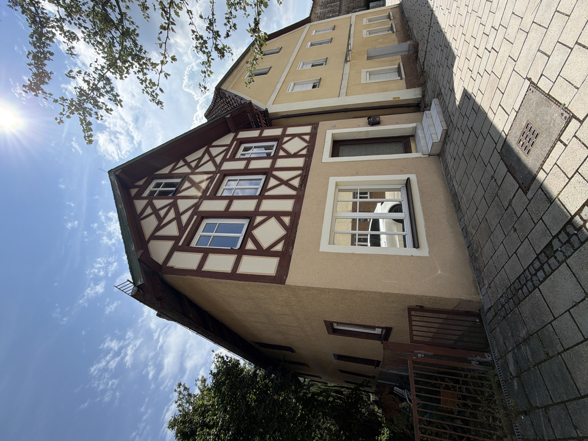 In Ebermannstadt, Germany, on a sunny day with scattered clouds, a traditional half-timbered building stands prominently on the left, its facade a mix of tan plaster and dark brown timber framing with diamond-shaped infills. The building features several windows, some with white frames. To its right, a yellow building with white trim runs parallel, also displaying multiple windows with white roller shutters. A paved walkway, made of grey rectangular stones and bordered by smaller dark stones, runs in front of both buildings. A rusty metal fence and dense green foliage are visible to the far left. Sunlight casts strong shadows across the walkway, illuminating a metal grate in the foreground.