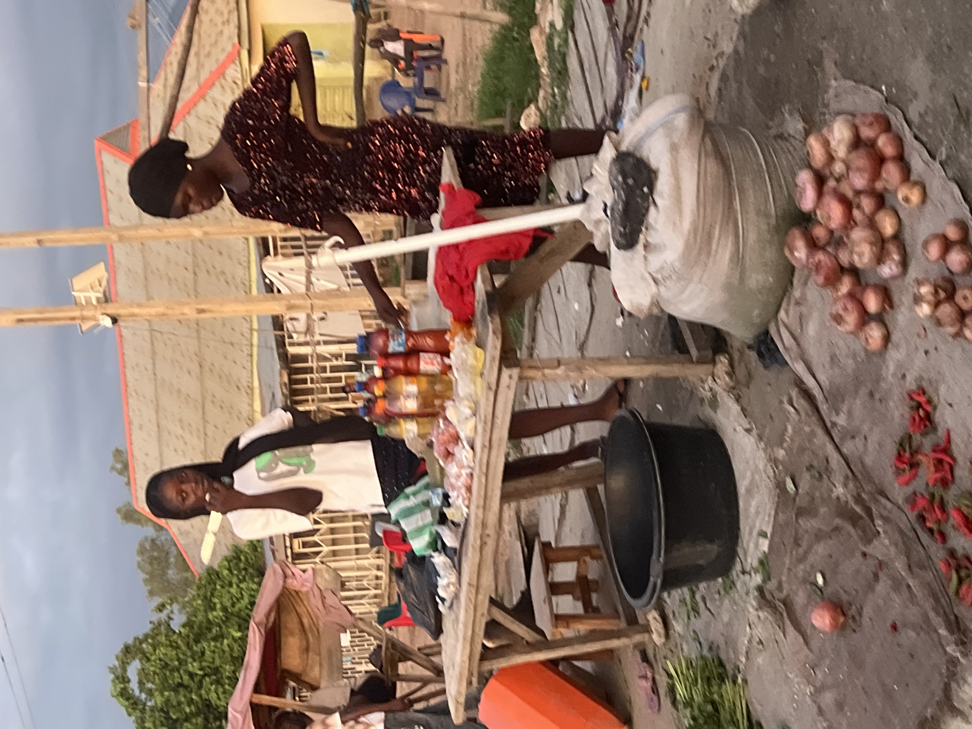 This image depicts an outdoor market scene in Howayi, Nigeria, likely during late afternoon or early evening under an overcast sky. Two women are central to the composition, standing behind and next to a wooden vendor's table.
The woman on the left wears a white short-sleeved t-shirt with a green graphic design, dark trousers, and has long dark hair. She holds a light-colored, elongated object to her mouth, appearing to be eating or chewing. Her gaze is directed slightly to the right of the viewer.
The woman on the right wears a dark red or maroon short-sleeved dress with a sequined or glittery texture, and a dark head covering. Her right hand rests on her hip, while her left hand is positioned near items on the table. She looks down towards the table.
The wooden table displays various items for sale, including multiple plastic bottles containing different liquids (possibly oils, sauces, or beverages), and several small packages of food items, some wrapped in clear plastic. A red cloth or garment is draped over the right side of the table. A green and white striped plastic bag is also visible on the table. Under the table on the left, a small wooden stool is present.
The ground in the foreground is uneven and appears to be dirt or concrete. A large black plastic basin or tub is positioned directly in front of the table. To the right of the basin, a large, light-colored sack, possibly filled with produce, is on the ground, topped with a dark, irregularly shaped object. Scattered around the sack and on the ground are piles of reddish-brown onions or shallots, and smaller piles of red chili peppers, some with green stems. A single, unpeeled onion lies to the left of the main onion pile. A small quantity of green leafy vegetables is visible on the far left ground. An orange plastic container is tilted on its side on the far left.
In the background, behind the vendors, are several structures, including houses with pitched roofs. One house has visible solar panels on its roof, and another features a yellow wall. Wooden poles and a metal fence or grille structure are also present. Further in the background, partial figures of individuals are visible, some seated on blue and red plastic chairs. Trees and other greenery are seen on the far left, under a muted, cloudy sky.