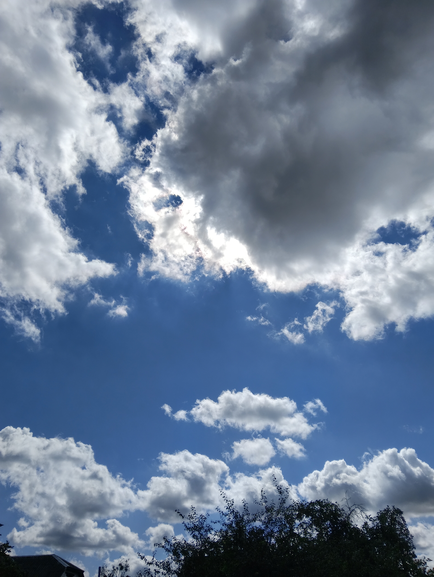 The image shows a sky filled with clouds over a cityscape. The sky is a deep blue, with scattered white and gray clouds. Some clouds are large and billowy, while others are thin and wispy. The sun is visible through a gap in the clouds, casting a bright light. In the foreground, the tops of trees and a building are visible, silhouetted against the sky. The scene appears to be set in the city of Hrodna, Belarus.