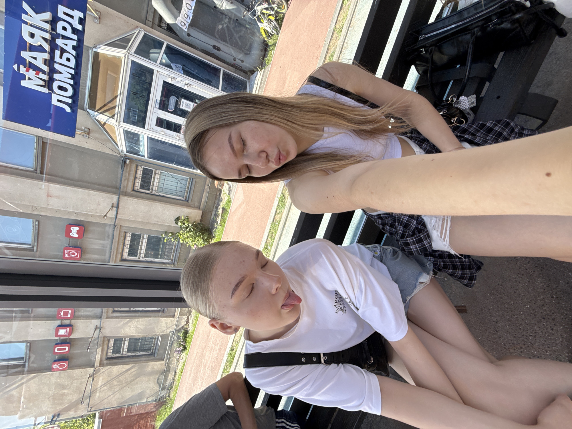 Two young women are sitting on a dark bench. The woman on the left is wearing a white t-shirt and denim shorts, with her tongue sticking out. She has a black purse slung over her shoulder. The woman on the right is wearing a white tank top and a plaid shirt over ripped denim shorts. She is puckering her lips. Behind them is a building with a blue sign that reads "МАЯК ЛОМБАРД" in white lettering. The building has several windows, some with bars. To the right, a yellow bicycle is visible. The scene appears to be outdoors, possibly in a city setting. The "МАЯК ЛОМБАРД" sign is a pawn shop chain with locations across Russia, including Perm.