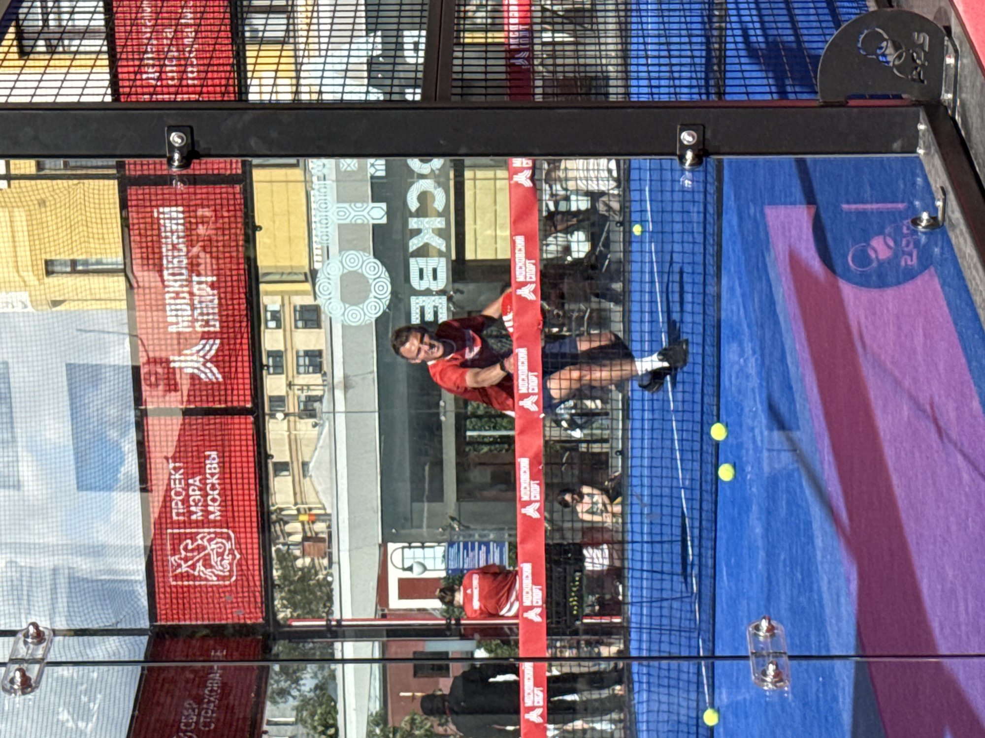This is a vertical shot of an outdoor padel tennis court with a man playing. The man, wearing a red shirt and dark shorts, is mid-action, hitting a yellow padel ball. The court has a blue surface with purple diagonal markings. A net divides the court.
The background is a blur of buildings and structures, suggesting an urban setting. Several banners with Cyrillic text are visible. One prominent red banner reads "ПРОЕКТ МЭРА МОСКВЫ" (Project of the Mayor of Moscow) along with a crest. Another banner has "МОСКОВСКИЙ СПОРТ" (Moscow Sport). The scene appears to be well-lit, indicating it's daytime, possibly late morning or afternoon, with sunshine casting shadows. The overall mood seems energetic, capturing a moment in a sports match. The metallic framework of the court and fences are visible, along with some glass panels reflecting the surroundings. Small yellow padel balls are scattered on the court.