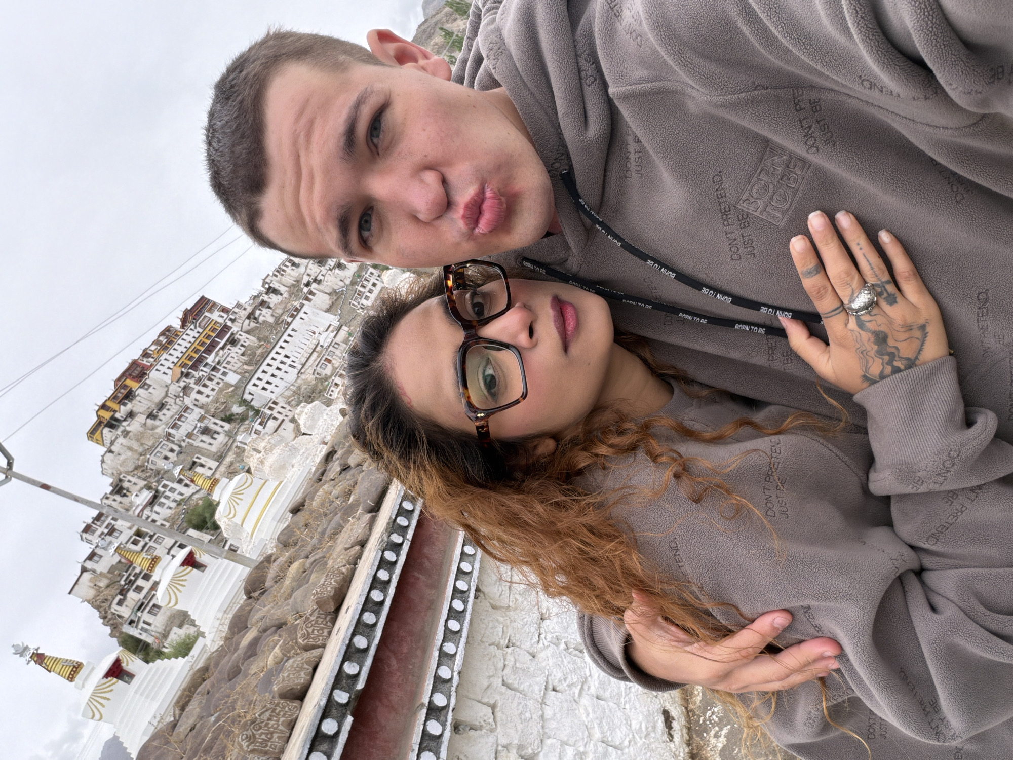 In the city of Thiksey, India, a young couple takes a selfie in front of a monastery. The woman, with curly auburn hair and large-framed glasses, leans into the man, who is puckering his lips for the camera. Both are wearing grey hoodies with repeating text. Behind them, the monastery complex cascades down a hillside, its white buildings and golden stupas stark against the cloudy sky. The foreground features a stone wall with decorative white circles and weathered rocks, some of which bear inscribed markings.