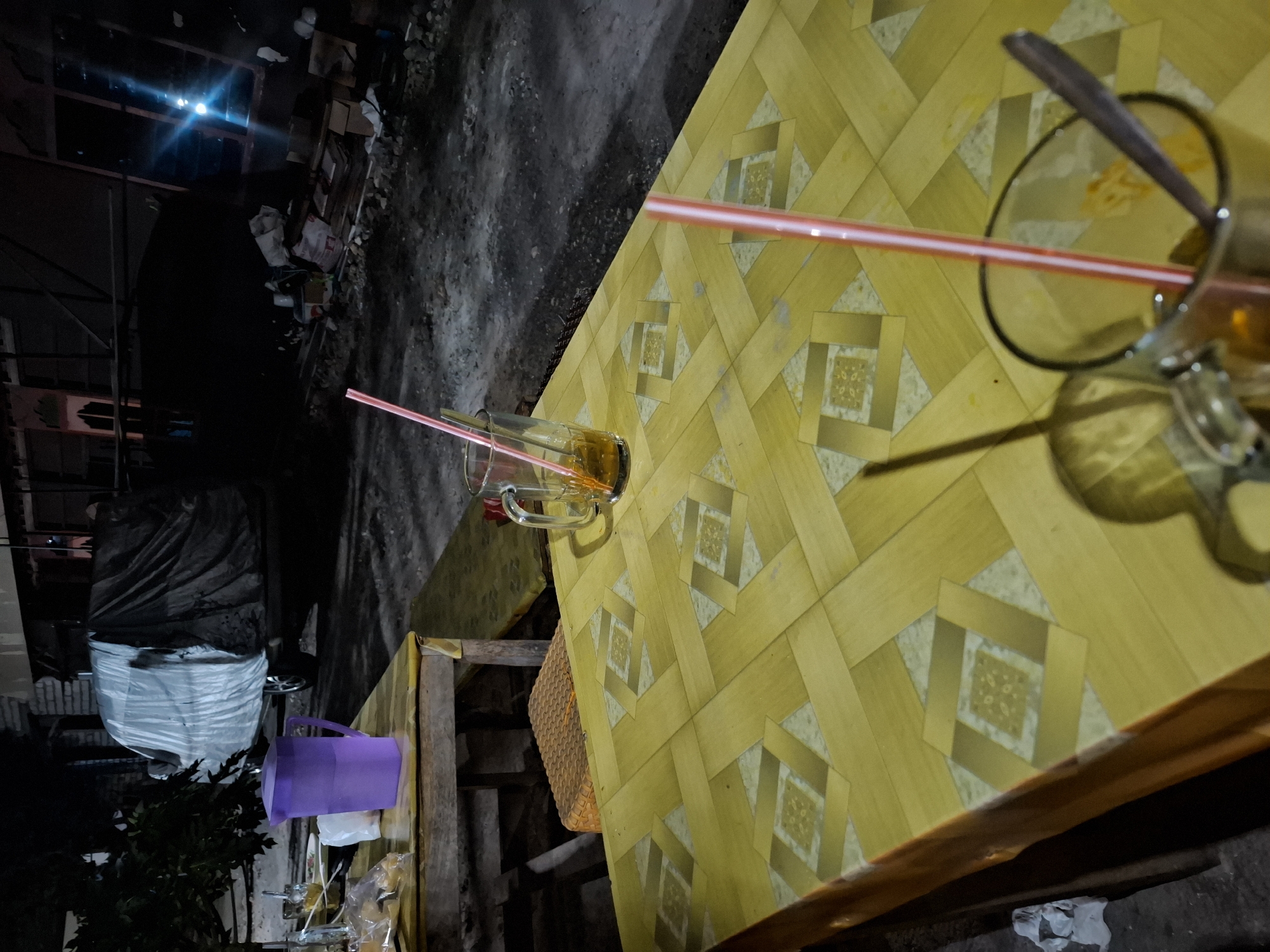 The image depicts an outdoor scene at night in Bulubrangsi, Indonesia, likely near a street or alley. In the foreground, a yellow table with a patterned surface is set with two glasses containing drinks and straws. One glass is a clear mug with a handle, filled with a brown liquid and a pink straw. Another glass, partially visible and out of focus, is also in the foreground with a red straw and a spoon. Behind the table, a purple pitcher is placed on a second, wooden table. To the left of the table, a portion of a tree with dark leaves is visible. In the background, a vehicle is covered by a large, dark tarp. Further back, there is a building structure with a window that emits a bright blue light. The ground appears to be dirt and gravel, with debris and piles of items scattered around. The overall atmosphere is dimly lit and appears to be a casual, outdoor setting, possibly a roadside eatery or a residential area at night.