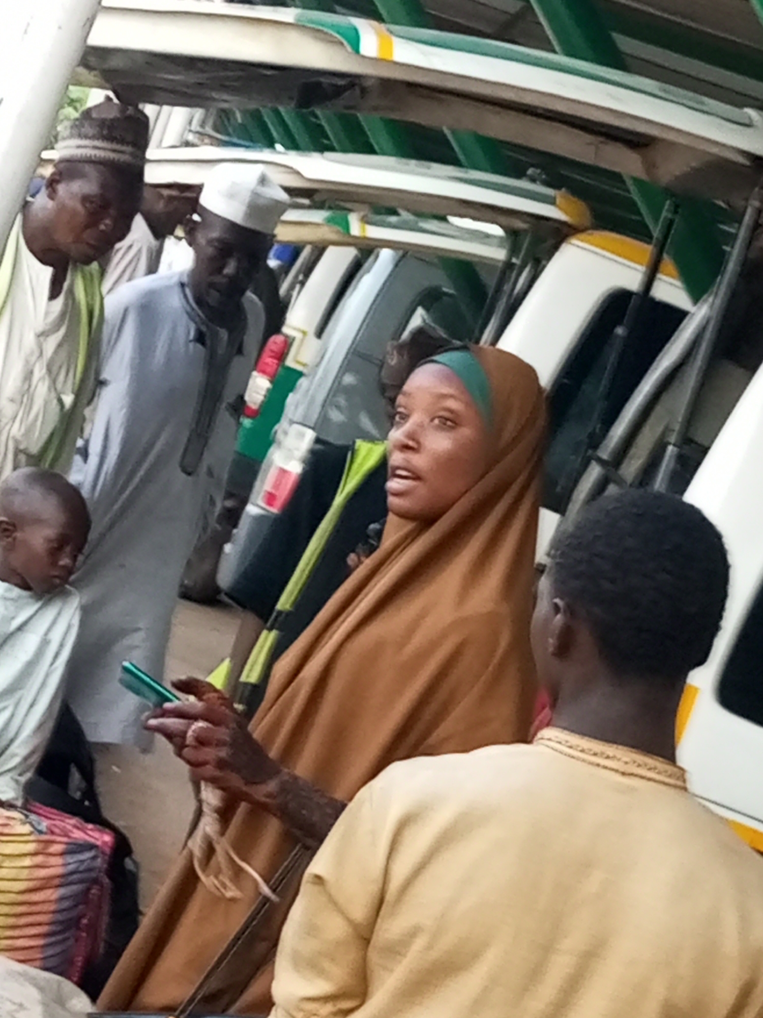 A group of people are gathered under a covered structure, likely a bus stop or market stall, in Gombe, Nigeria. The environment appears to be outdoors, with a natural light source illuminating the scene. In the foreground, a woman wearing a brown hijab and green headscarf is holding a cell phone. Her hand, adorned with henna, is visible as she interacts with the device. To her right, a young person in a light yellow garment, facing away from the camera, is also present. Behind the woman, a boy in a light blue garment is looking down. Further back, two men are visible. One man, wearing a white cap and light blue traditional attire, is looking towards the right. To his left, another man in a patterned cap and light colored clothing, also looking to the right. Several vehicles, possibly buses or minibuses, are parked behind the people, suggested by their white and yellow exteriors and tinted windows. The overall scene depicts a moment of everyday life in Gombe, with individuals engaged in various activities.