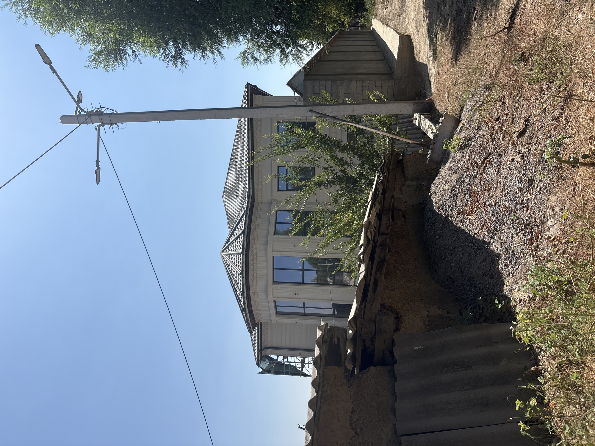 The image depicts a residential street in Shymkent, Kazakhstan, on a clear, sunny day. In the foreground, a pile of gravel and construction debris is visible, with weathered corrugated metal sheets leaning against a low, crumbling wall. To the right of this is a dirt path leading towards a modern, two-story house with a distinctive multi-faceted roof and large windows. A concrete utility pole with multiple streetlights and electrical wires stands prominently in the middle ground, casting a shadow. Adjacent to the house is a long, low-profile concrete structure. The background is dominated by a clear blue sky and lush green trees, suggesting a suburban or semi-rural setting within the city. No people are present in the scene, and the focus is on the architectural elements and the surrounding environment.