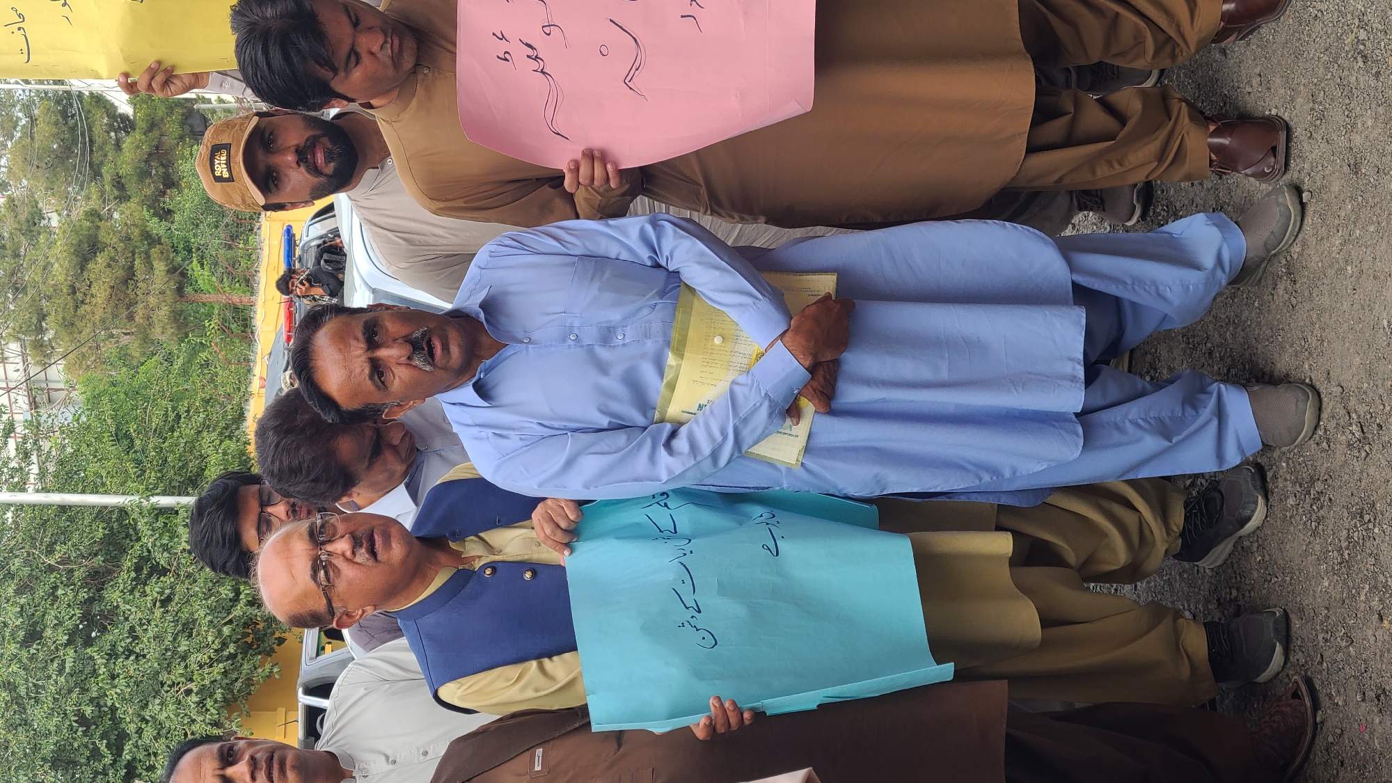 A group of men stand in a row on an asphalt surface, possibly a street or parking lot, holding signs with writing in Urdu. The man in the center wears a light blue shalwar kameez and holds a yellow folder. The man in front of him wears a yellow shalwar kameez with a blue vest and holds a blue sign. The other men wear various shades of brown shalwar kameez and hold pink and yellow signs. In the background are trees, a utility pole, and a car. The image appears to be taken during a protest or demonstration. The location is likely in or near Quetta City Tehsil, Pakistan.