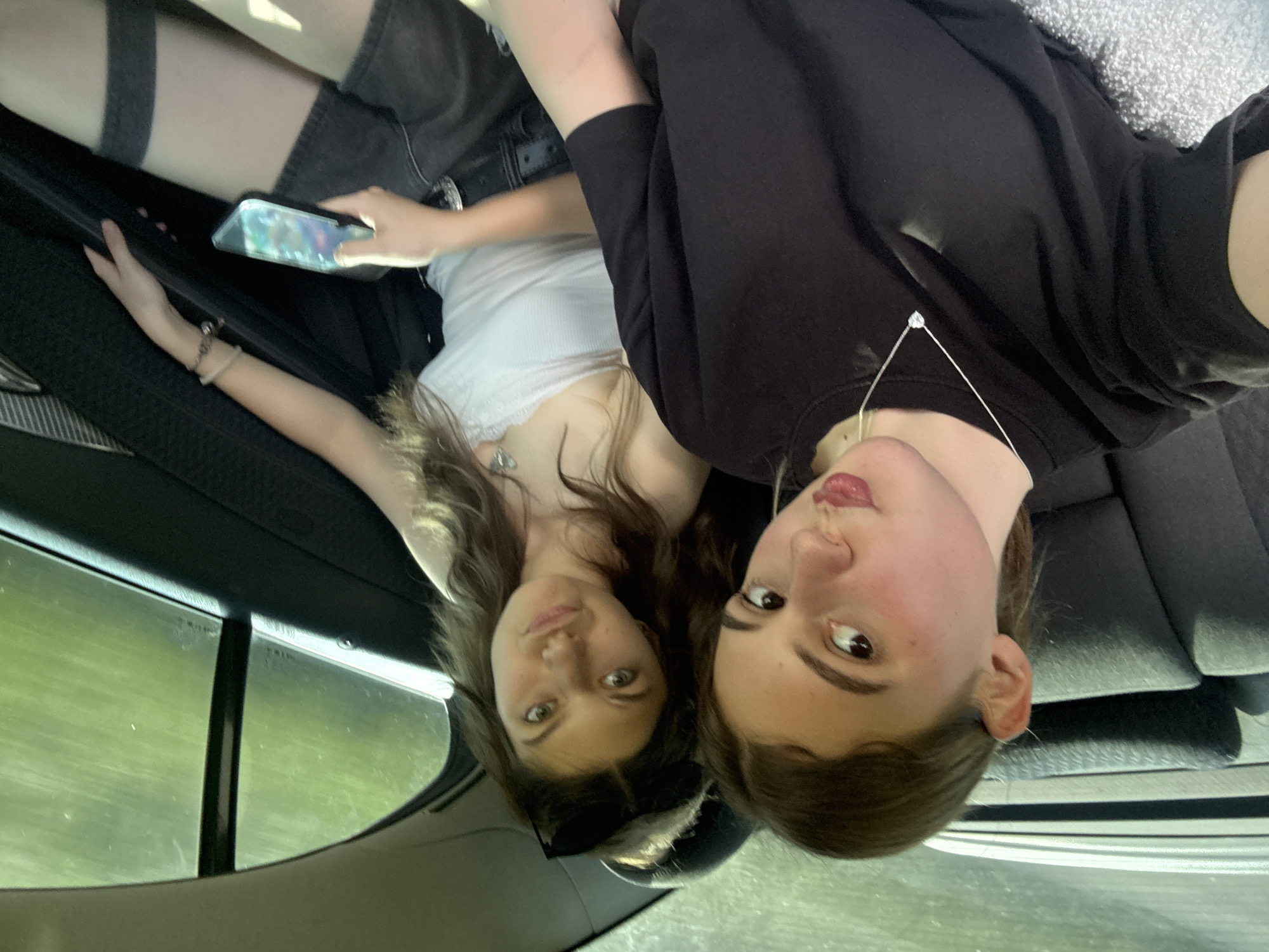 Two young women are in the back seat of a car, taking a selfie. The woman on the left has long brown hair and is wearing a white tank top and black shorts with thigh-high socks. She is holding a smartphone in her hand. The woman on the right has shorter brown hair and is wearing a black t-shirt with a silver pendant necklace. Both women are looking at the camera. The car's interior is visible, with dark seats and a window showing a blurry green landscape, suggesting they are driving through a rural area, possibly near Koprivshtitsa, Bulgaria, given the location context. The lighting suggests it's daytime, perhaps late afternoon due to the soft glow. The women appear relaxed and are sharing a casual moment. There is no visible text in the image.
