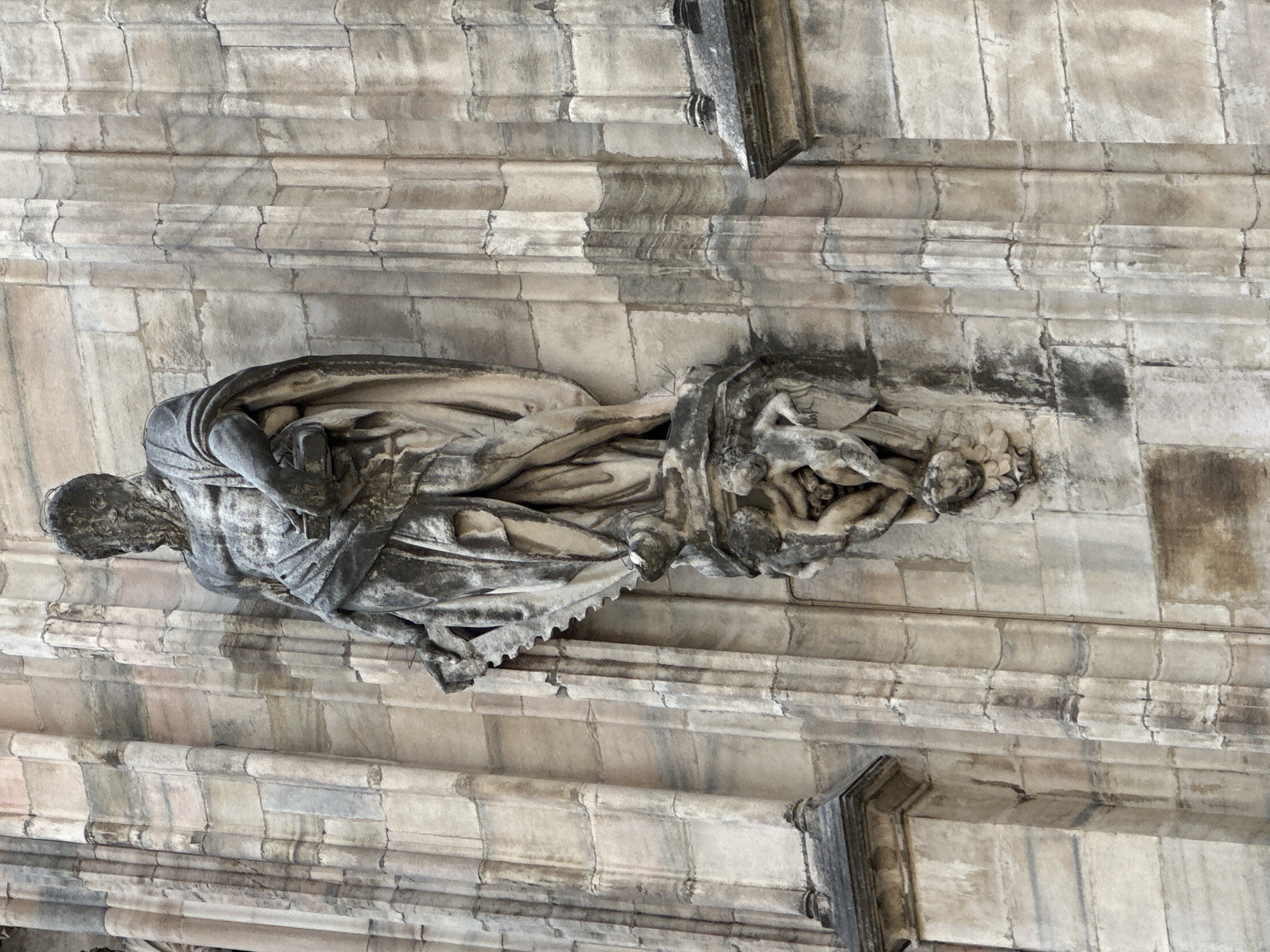 The image shows a detail of a stone sculpture on the exterior of the Milan Cathedral (Duomo di Milano) in Milan, Italy. The sculpture features a bearded male figure wearing a toga and holding a book or tablet in his left hand. Below the main figure, there is a smaller sculpture of cherubic figures. The stone appears weathered and aged, typical of the centuries-old architecture of the Duomo. The background is a section of the cathedral's facade, characterized by its intricate stonework and Gothic architectural elements. The lighting suggests it is daytime, with sunlight highlighting the textures of the stone.