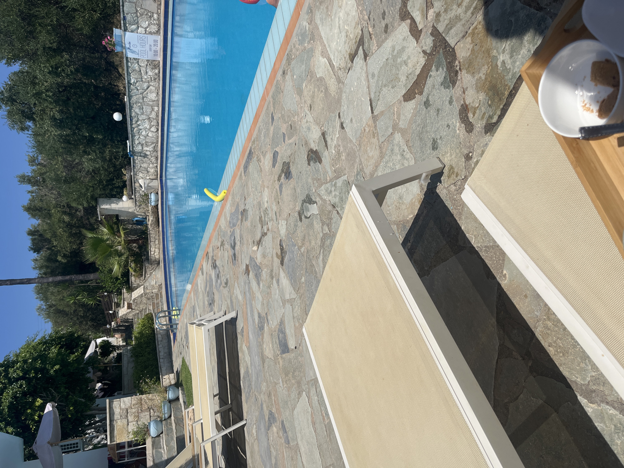 The image shows a poolside area at the resort of Exopoli, Greece. In the foreground, two empty beige lounge chairs are positioned on a stone patio. The pool itself is a bright blue, with a yellow pool noodle floating near the edge. A stone wall with a sign indicating "POOL HOURS 10:00 - 20:00" is visible behind the pool, surrounded by lush green trees and vegetation. The sun is shining brightly, casting shadows on the patio. There are no people visible in the immediate vicinity of the pool.