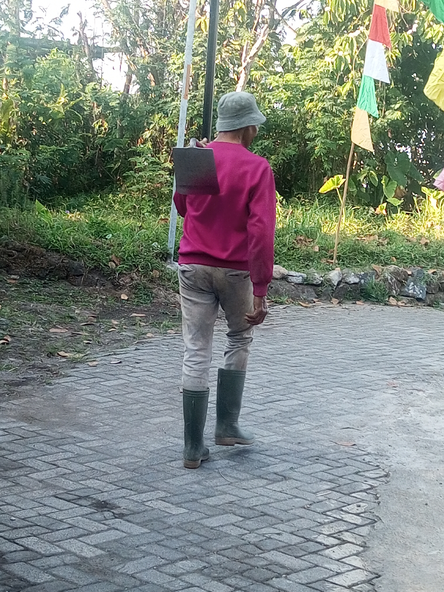 A person wearing a faded green bucket hat, a bright magenta long-sleeved shirt, light brown trousers, and dark green rubber boots stands facing away from the camera. They are carrying a rectangular, dark-colored tool with a metal head and a handle over their right shoulder. The person is walking on a paved walkway composed of interlocking gray rectangular bricks laid in a herringbone pattern. To the right of the walkway, there is a patch of wet concrete. The background features lush green foliage, including trees and grass, with a low stone wall visible in the middle ground. Several colorful triangular flags—red, white, green, and yellow—are also visible on poles to the right of the frame, set against the greenery. The scene appears to be outdoors, likely in a rural or semi-rural setting in Andongsili, Indonesia, given the vegetation and flags often associated with local celebrations or events. The lighting suggests it is daytime, with sunlight filtering through the trees.
