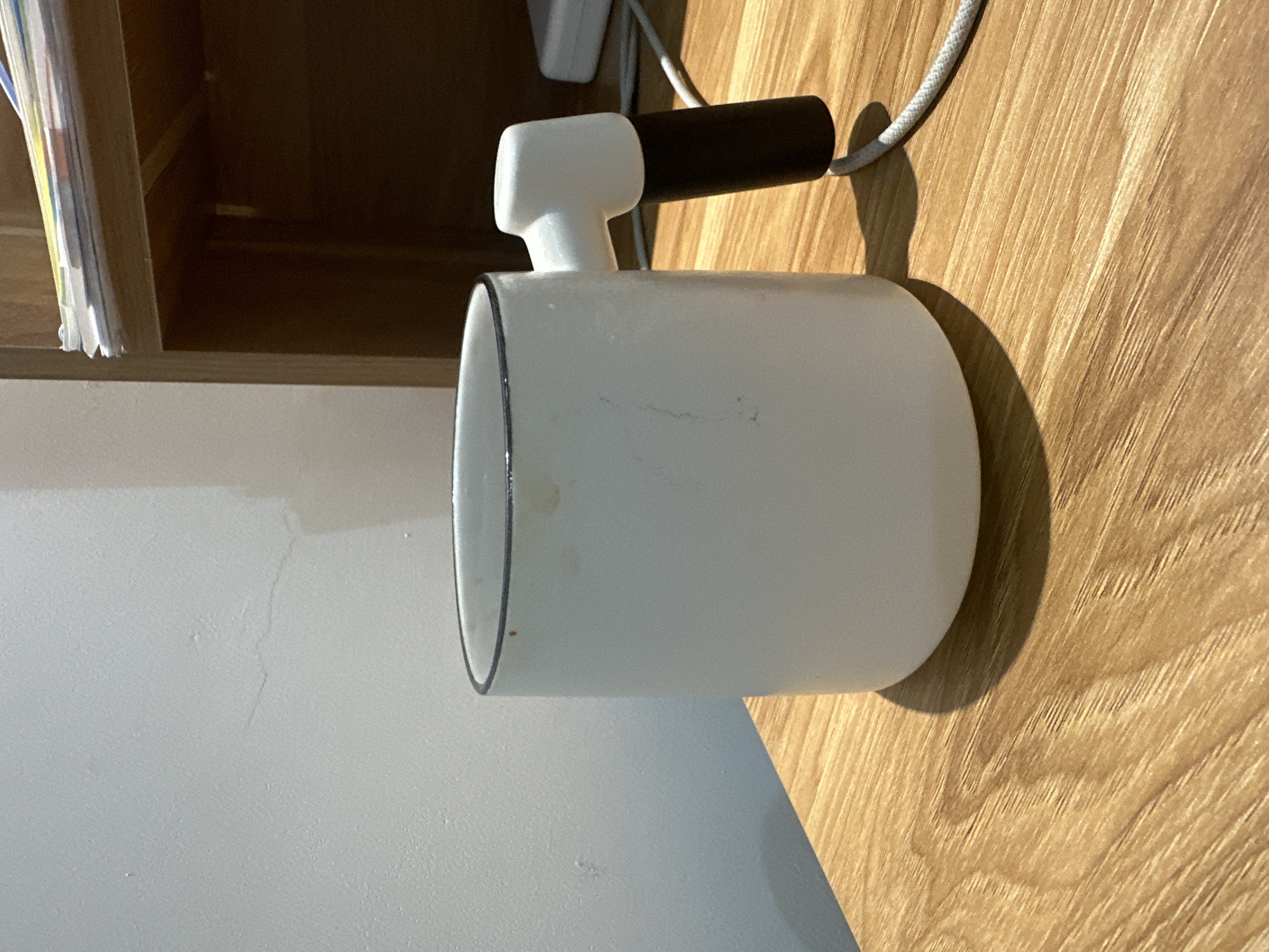 A white cylindrical lamp with a black handle and a cord is positioned on a wooden surface next to a white countertop. The lamp is casting a shadow on the wood. A bookshelf is partially visible in the background. The scene is indoors and appears to be a domestic environment. There are no people present. The location is unspecified beyond that this is somewhere in the Bac Nha Trang Ward, Vietnam.