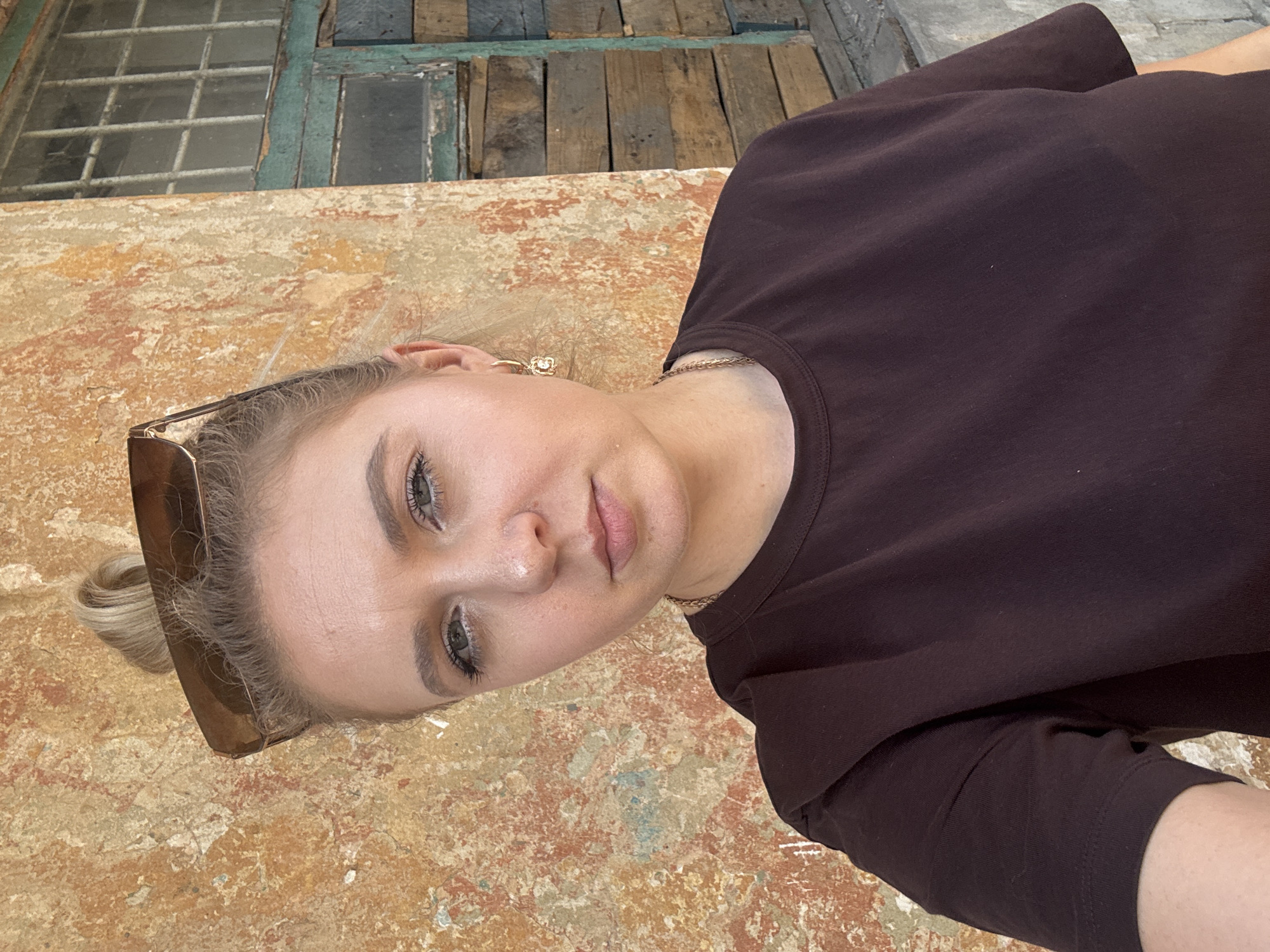 A young woman with blonde hair styled in a bun, wearing sunglasses perched on her head and a dark brown t-shirt, stands in front of a textured, weathered wall. She is looking directly at the camera with a neutral expression. To her right, part of an old window frame with peeling green paint and wooden shutters is visible. The overall environment appears to be outdoors, possibly in an urban setting given the architectural elements.