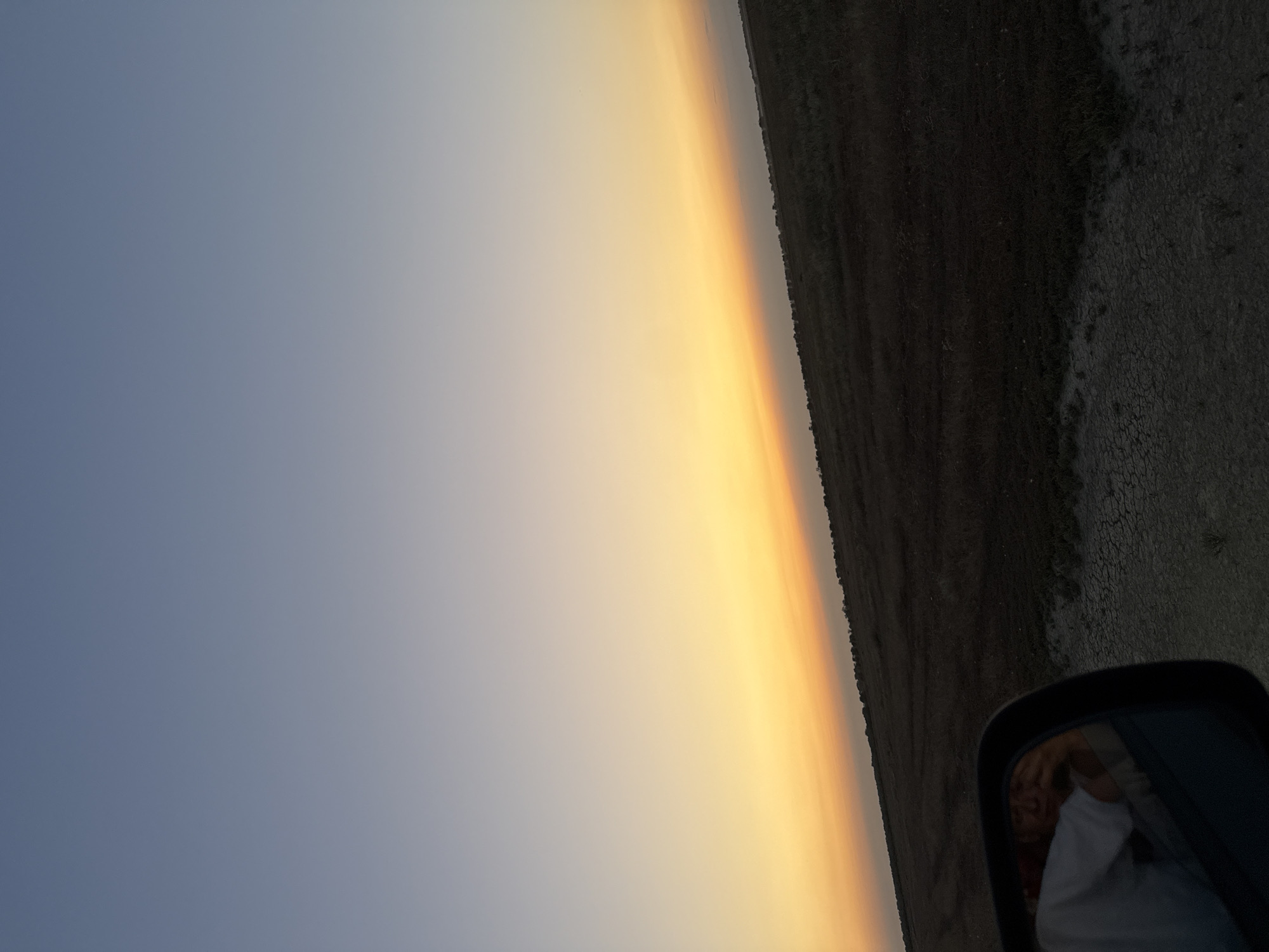 The image captures a sunset scene with a gradient sky transitioning from a pale blue at the top to warm orange and yellow hues near the horizon. A dark, flat landscape with sparse vegetation stretches across the lower portion of the frame. In the bottom left corner, a car's side mirror is visible, reflecting a person wearing a white shirt, their face obscured as they appear to be taking the photograph. The foreground shows a dirt or gravel surface, possibly a road. The location is identified as Урожайновское сельское поселение, Ukraine.