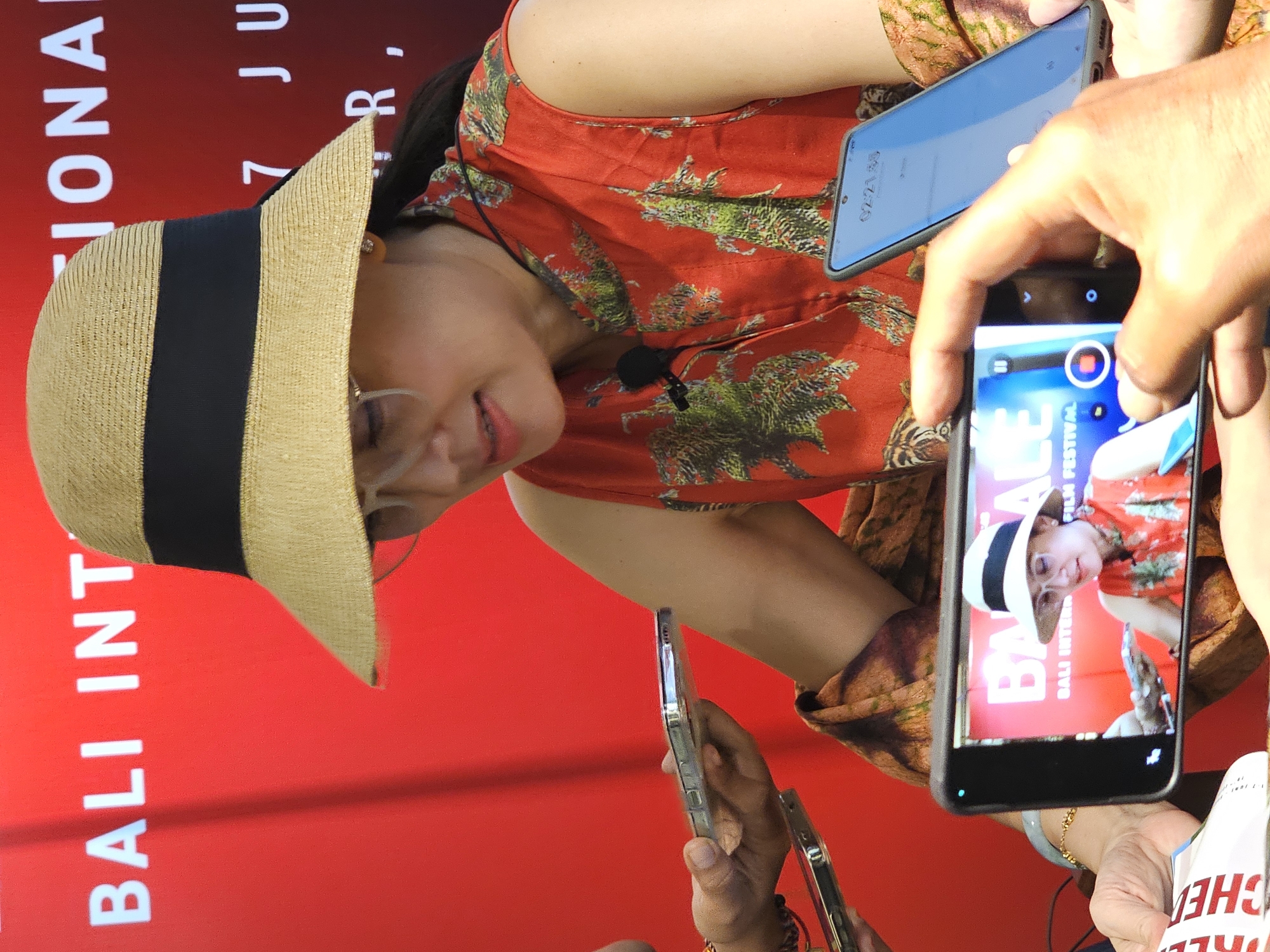 This close-up image captures a woman at what appears to be a film festival or public event in Denpasar, Indonesia. The woman is smiling and wearing a straw hat with a black band, a red top with green foliage patterns, and glasses. A small microphone is clipped to her top. She is the central subject, being actively photographed or filmed by multiple individuals using smartphones.
Several hands are visible holding these devices, with one prominent smartphone in the foreground clearly displaying a video recording interface and reflecting the scene, including the woman and the red background. On its screen, the text "BAALE BALI INTER FILM FESTIVAL" is distinctly visible, confirming the event type and location. Another phone, held by the woman herself, shows "02:21.38" which appears to be a digital clock or timer.
The background consists of a large, vibrant red banner or screen with white text. Partially visible text on the banner includes "BALI INT" (likely part of "BALI INTERNATIONAL"), "JONA", "7 JU" (suggesting "7 JULY"), and "R,". A partial word "CHE" is also seen in the lower right corner. The lighting suggests a bright setting, possibly indoors or under an awning during daytime. The overall mood conveyed is one of engagement and activity, centered around the documented subject.