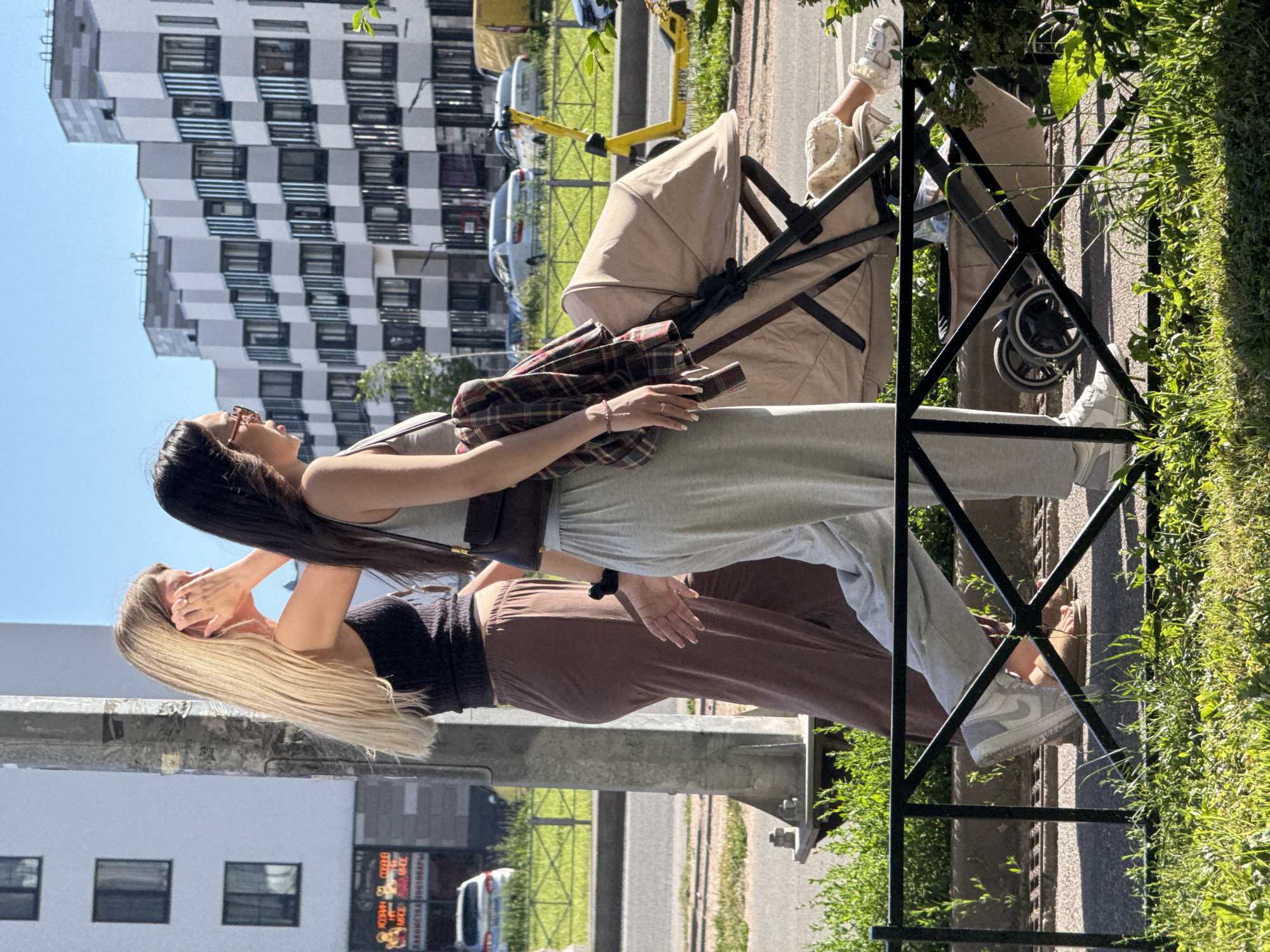 Two women are walking side-by-side on a sidewalk in Saint Petersburg, Russia. The woman on the left has long blonde hair and is wearing a black tank top and brown wide-leg pants. She has her right hand raised to her ear, and she is wearing a ring on her right index finger. The woman on the right has dark hair and is wearing a light gray tank top and light gray sweatpants. She has a plaid shirt draped over her left arm and is wearing a brown crossbody bag. She has a ring on her left ring finger. Between them, they are pushing a beige stroller. A child's legs, clad in white socks, are visible inside the stroller. The background features a modern apartment building with geometric balconies and a blue sky. A yellow electric scooter is parked on the sidewalk across the street, and several cars are visible in the distance. The foreground shows a black metal fence and green grass. The scene appears to be a sunny day.