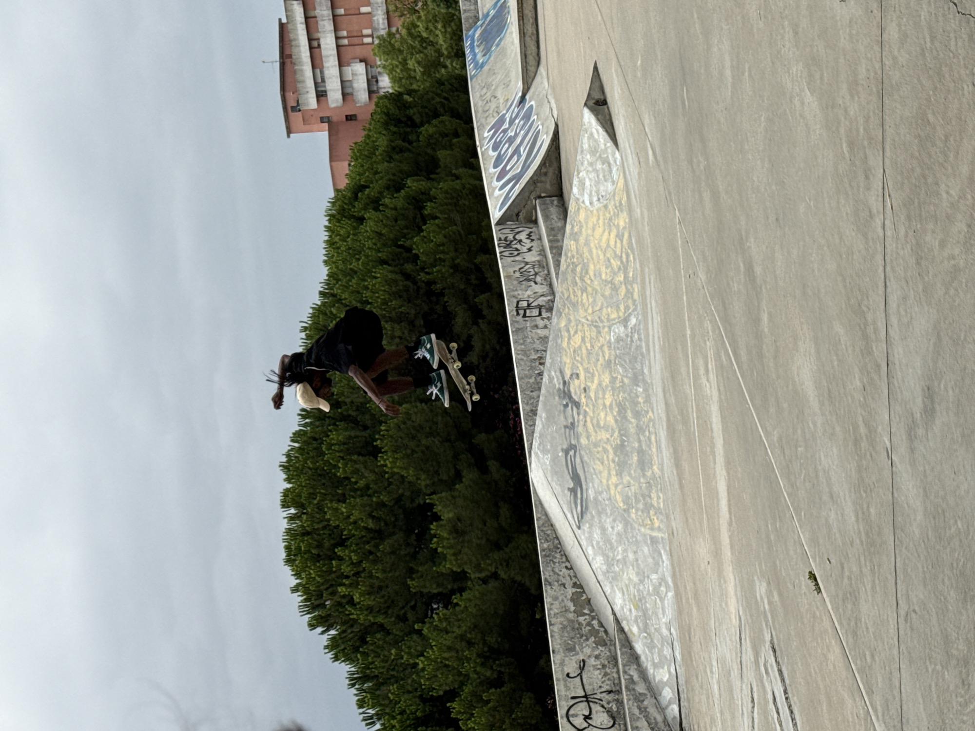 A young Black person wearing a black t-shirt, black shorts, a tan baseball cap, and green and white sneakers is captured mid-air performing a skateboard trick in a skate park. The skateboarder is positioned above concrete ramps, with their body angled as if executing a jump. Their arms are extended, one reaching towards their head and the other towards the skateboard, suggesting an active movement. The skateboard itself is visible beneath their feet.
The skate park features various concrete structures, including ramps and ledges, some adorned with graffiti in shades of black, blue, and purple. In the background, a dense line of dark green trees forms a natural backdrop. Behind the trees, a multi-story building with a reddish-pink facade and white window frames stands against an overcast sky. The overall lighting suggests a cloudy day.
The skate park is likely located in Almada, Portugal. The architectural style of the building and the type of vegetation are consistent with the region.