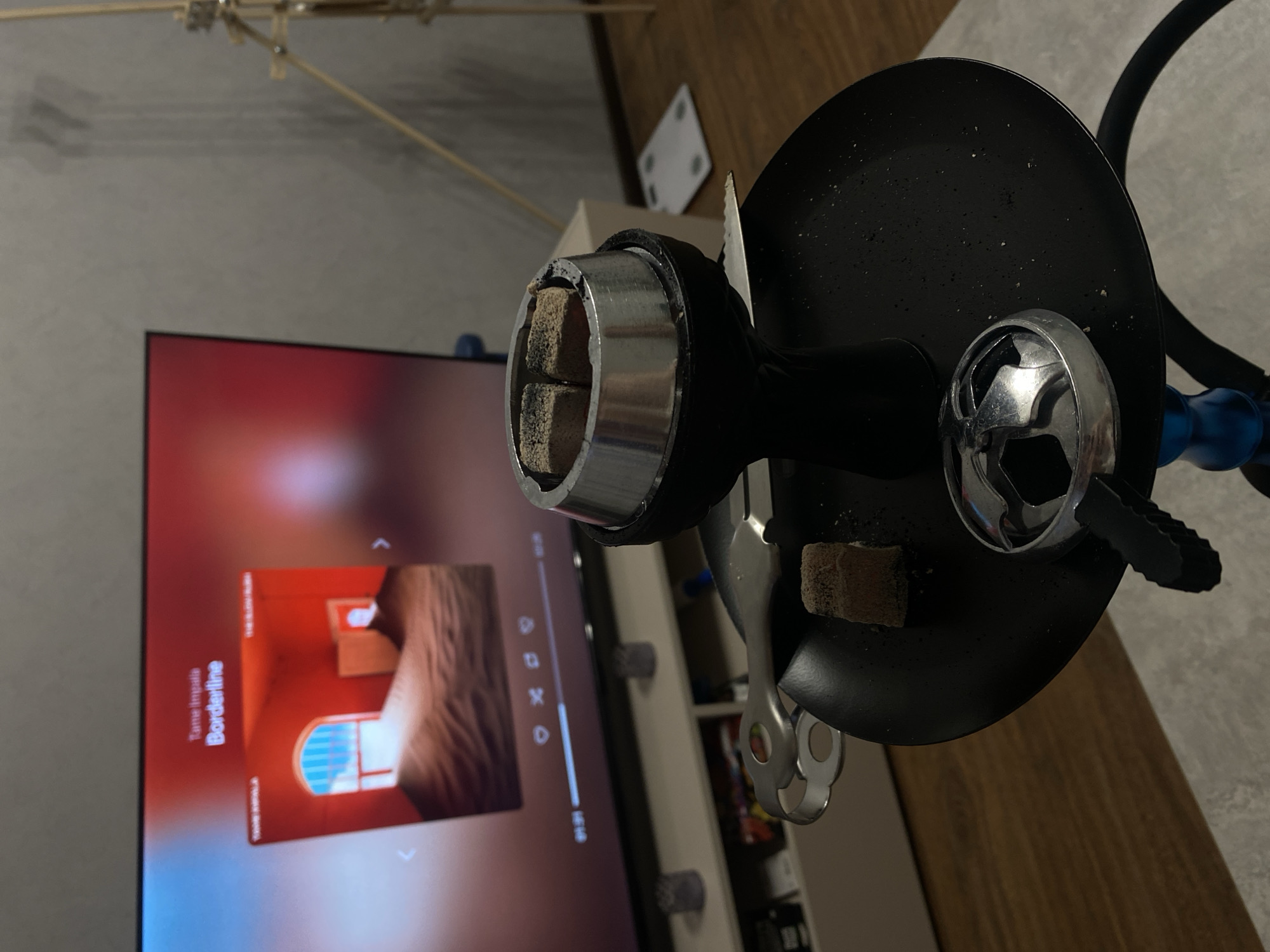 The image shows a hookah, a traditional water pipe, with lit coals on top, indicating active use. The hookah is black with silver accents, placed in front of a television screen. The TV is displaying an image with the song "Borderline" by Tame Impala displayed at the bottom left. The environment is a room with a shelf behind the TV. There is no evidence to pinpoint this location to Izhevsk, Russia, or any other specific city. No people are visible.