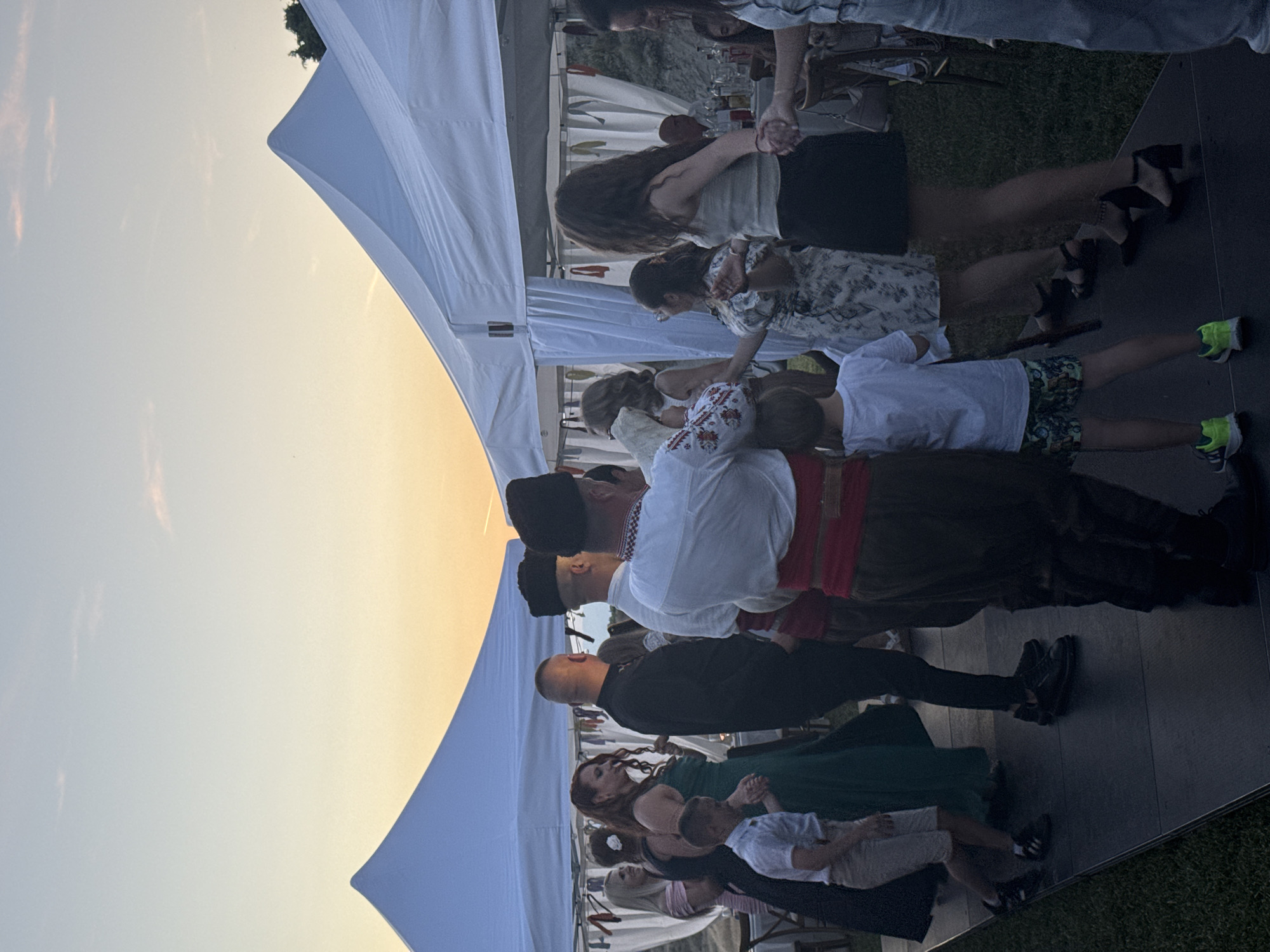 An outdoor event is captured at dusk in Harsovo, Bulgaria, featuring a gathering of individuals under two white, peaked event tents. The sky transitions from a pale blue at the top to a warm orange-yellow near the horizon, with faint, wispy clouds visible. A dark, modular dance floor is set up on a manicured green lawn.
On the dance floor, multiple individuals are engaged in dancing. On the left, a woman with long, reddish-brown hair, wearing a dark green dress, holds hands with a young boy in a white polo shirt and light shorts. A man in dark clothing stands facing right, next to them. Behind these individuals, a woman with blonde hair in a light-colored dress is partially visible, smiling, and another woman in a dark dress is also partially visible. Under the tent on the left, white drapes and tables with chairs are discernible in the background.
Towards the center and right, several men are visible from behind, dressed in traditional attire consisting of white shirts with red and white embroidery on the sleeves, dark pants or trousers, and distinctive black hats. At least two of these men wear wide red sashes around their waists and appear to be participating in a coordinated dance. A second young boy in a white t-shirt and patterned shorts stands between two of these traditionally dressed men, facing right. Further right, a woman with long, dark, curly hair, wearing a light-colored top and dark skirt, is dancing and holding hands with other individuals. Another woman in a patterned dress is dancing nearby. In the background under the right tent, a man sits at a table with bottles. Footwear includes dress shoes, sneakers, and heels. The tents are adorned with white fabric drapes and subtle decorative elements.