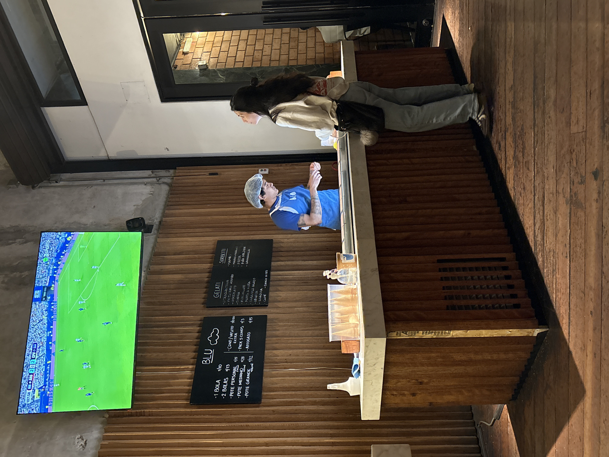 In San Isidro, Peru, at an establishment that appears to be a gelato shop, a female customer stands at a counter, looking upwards. She is wearing light-colored jeans, a cream-colored long-sleeved shirt, and a dark shoulder bag with a fluffy pom-pom. She is facing a male employee who is behind the counter, wearing a blue t-shirt and a hairnet. The employee is in the process of serving the customer, with his hands near the counter.
The counter is made of wood and topped with a light-colored surface. On the counter, there are several ice cream cones in a clear plastic container, a spray bottle, a bottle of hand sanitizer, and a small potted plant or container. Behind the counter, a wooden wall is decorated with chalkboards displaying prices and menu items for "BLU GELATI" and "SORBETTI".
Above the counter, a large flat-screen television displays a live soccer match. The room has a rustic ambiance with exposed concrete walls and a wooden floor. The overall lighting suggests it is evening or indoors with artificial lighting.
