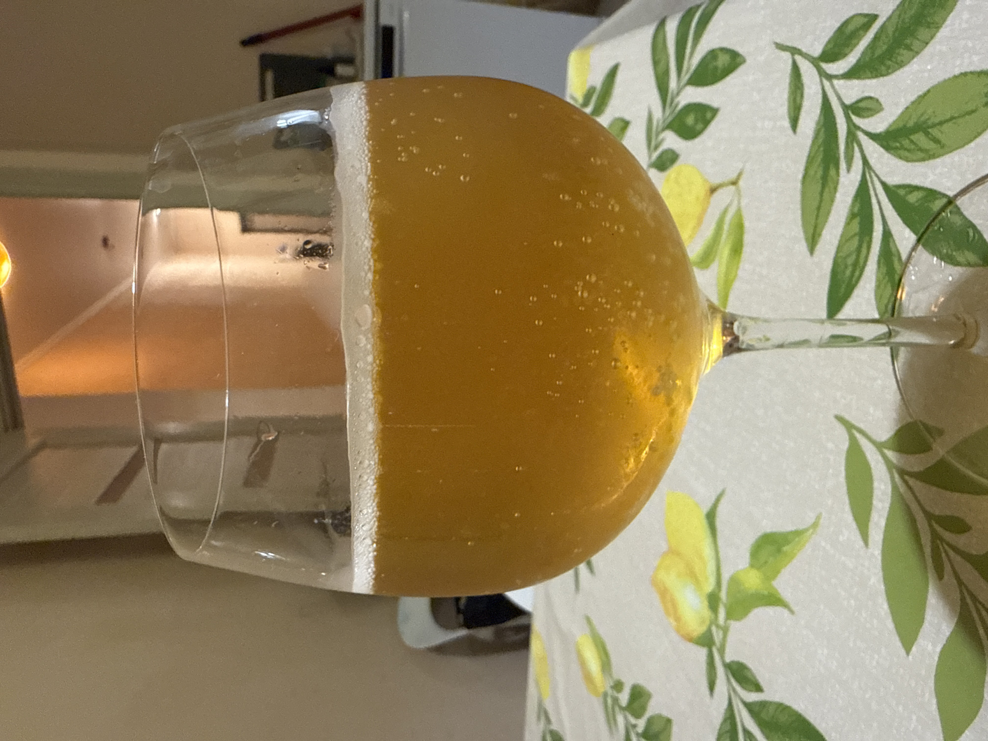A close-up, rotated photograph captures a stemmed glass, likely a wine glass or flute, filled with a golden, effervescent beverage. The drink, possibly beer or champagne, has a significant head of white, frothy foam at the top, and numerous small bubbles are visible throughout the liquid and clinging to the inside of the glass.
The glass is placed on a table covered with a tablecloth that features a bright pattern of yellow lemons and green leaves against a light, possibly white or cream, background. A portion of the glass's stem and base are visible, extending towards the right side of the frame.
The background is softly blurred, indicating an indoor setting with light-colored walls or surfaces and indistinct furniture or appliances. A warm, bright light source casts a soft glow in the upper left background. The overall focus is on the sparkling drink and its presentation on the patterned table setting. There are no people visible, and no text can be deciphered.
