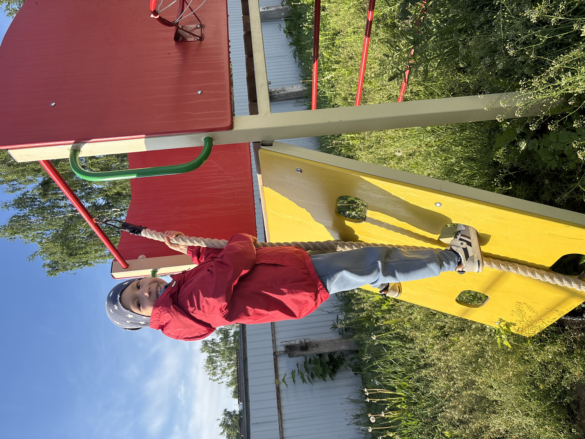 A young child is pictured playing on a brightly colored playground structure outdoors in Chaikovsky, Russia. The child, likely a boy, is wearing a red jacket, light blue pants, a blue and white star-patterned hat, and sneakers. He is actively engaged, holding onto a thick, twisted white rope that forms part of the climbing apparatus, and appears to be suspended or climbing on it. A cheerful smile is visible on his face as he looks towards the camera.
The playground equipment consists of large red and yellow panels, with green and red metal bars and supports. One green curved handle is visible near the top left of the frame. The rope the child is holding stretches from a red part of the structure down towards a yellow climbing board.
The setting is bright and sunny, indicating clear weather, likely during the morning or late afternoon given the strong shadows. In the background, there are lush green grasses and various wild plants, including some with small white flowers and dandelions. Trees with green foliage are visible against a clear, expansive blue sky with a few faint wispy clouds. Towards the middle and right of the background, an industrial-looking corrugated metal structure or fence is partially visible. No visible text is present in the image.