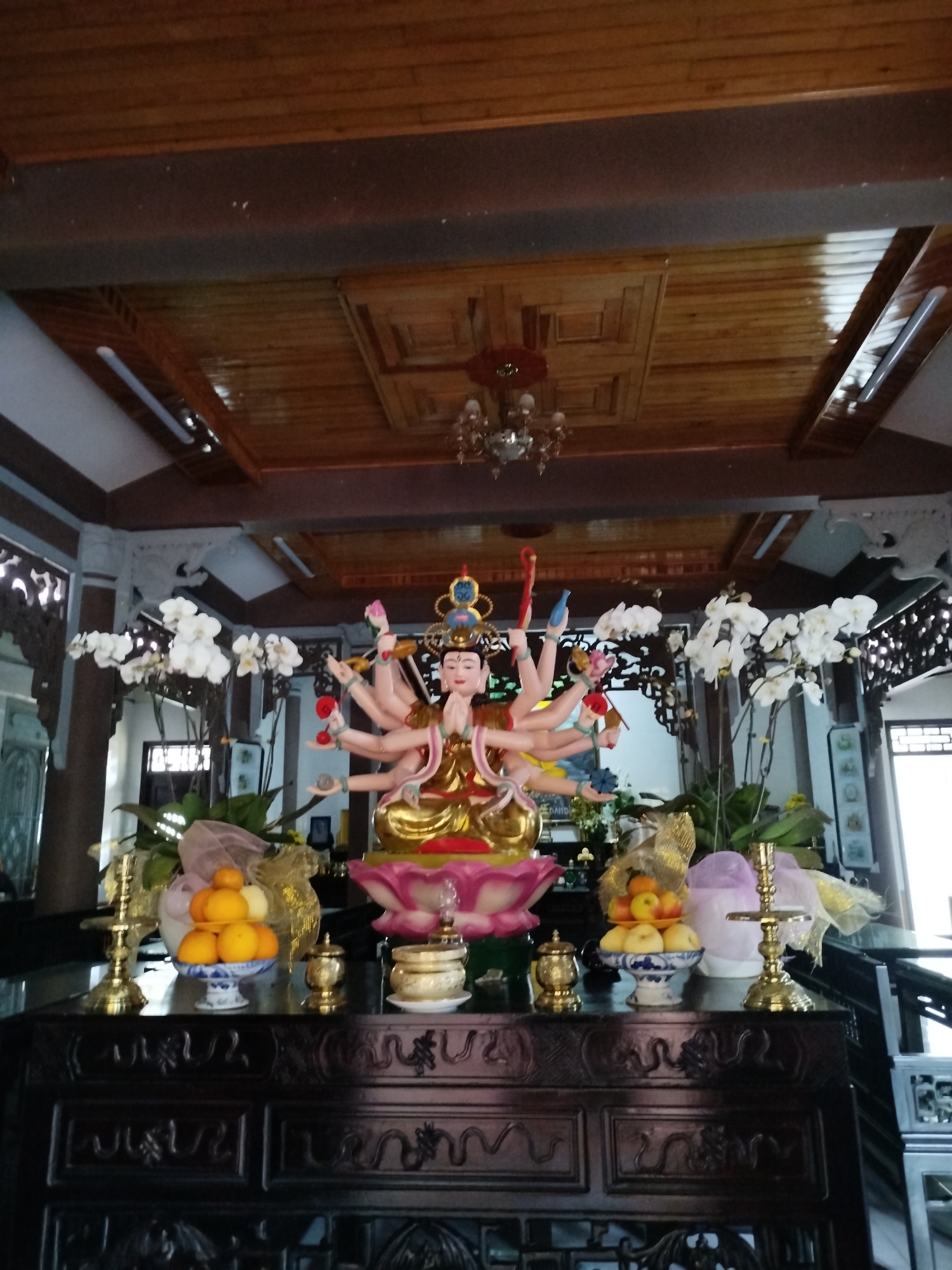 The image shows a shrine in a room with a wooden ceiling, likely located in a home or temple. The main focal point is a statue of a deity with multiple arms, seated on a lotus flower base. The statue is adorned with golden garments and a jeweled crown. Orchids, in full bloom, flank the statue on either side. In front of the statue are offerings of fruit, including oranges and apples, arranged in porcelain bowls. Gilded candleholders and other ornate containers are also present. The shrine is placed on a dark, intricately carved wooden table or altar. From the context provided (Xuân Trường - Đà Lạt, Vietnam), this could potentially be a Buddhist or folk religion shrine, as these are common in the region. The setting is peaceful and reverent.