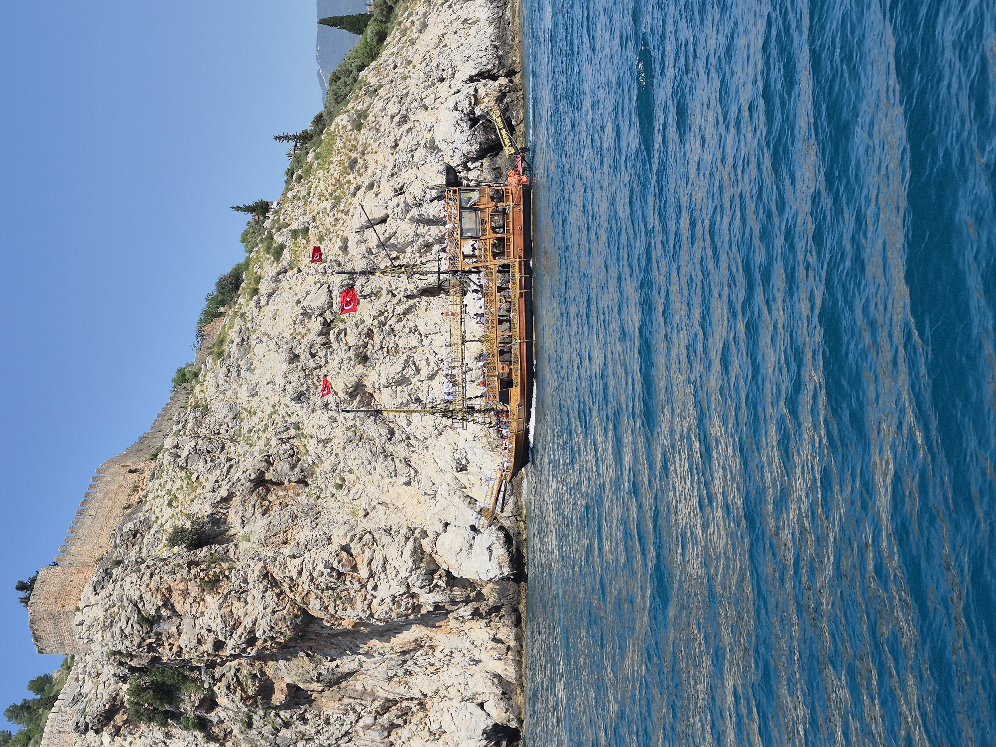 The image depicts a tourist boat docked along the rocky coast of Alanya, Turkey, characterized by a steep, light-colored cliff face with sparse vegetation. The cliff is topped by a medieval castle wall. Several Turkish flags are displayed along the cliff and on the boat. The boat is multi-decked with a yellow railing and several people are visible onboard. To the right of the boat is the bright blue water of the Mediterranean Sea, with visible wave patterns. The sky is clear and light blue.
