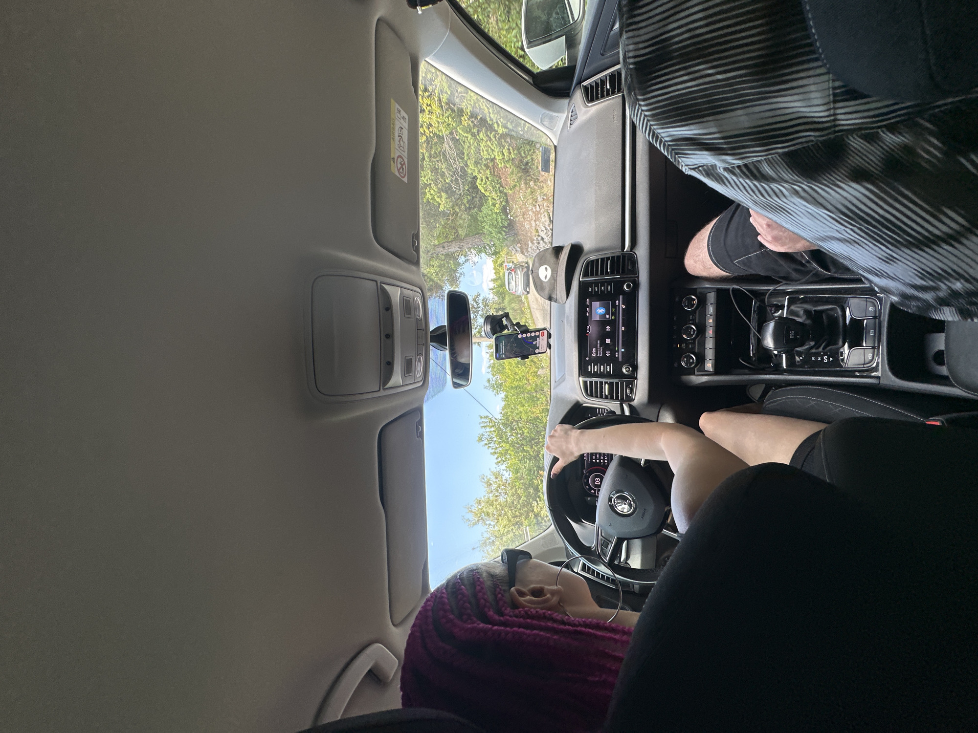 A view from inside a car, likely the driver's seat, shows a person with pink braided hair driving the car. The person is wearing glasses. The dashboard and steering wheel are visible. Outside the windshield, a road and trees are visible, suggesting a drive in a rural or forested area. A passenger sits in the front passenger seat. Based on the location information, the scene is likely in Kubasi, Montenegro, characterized by its natural landscape.