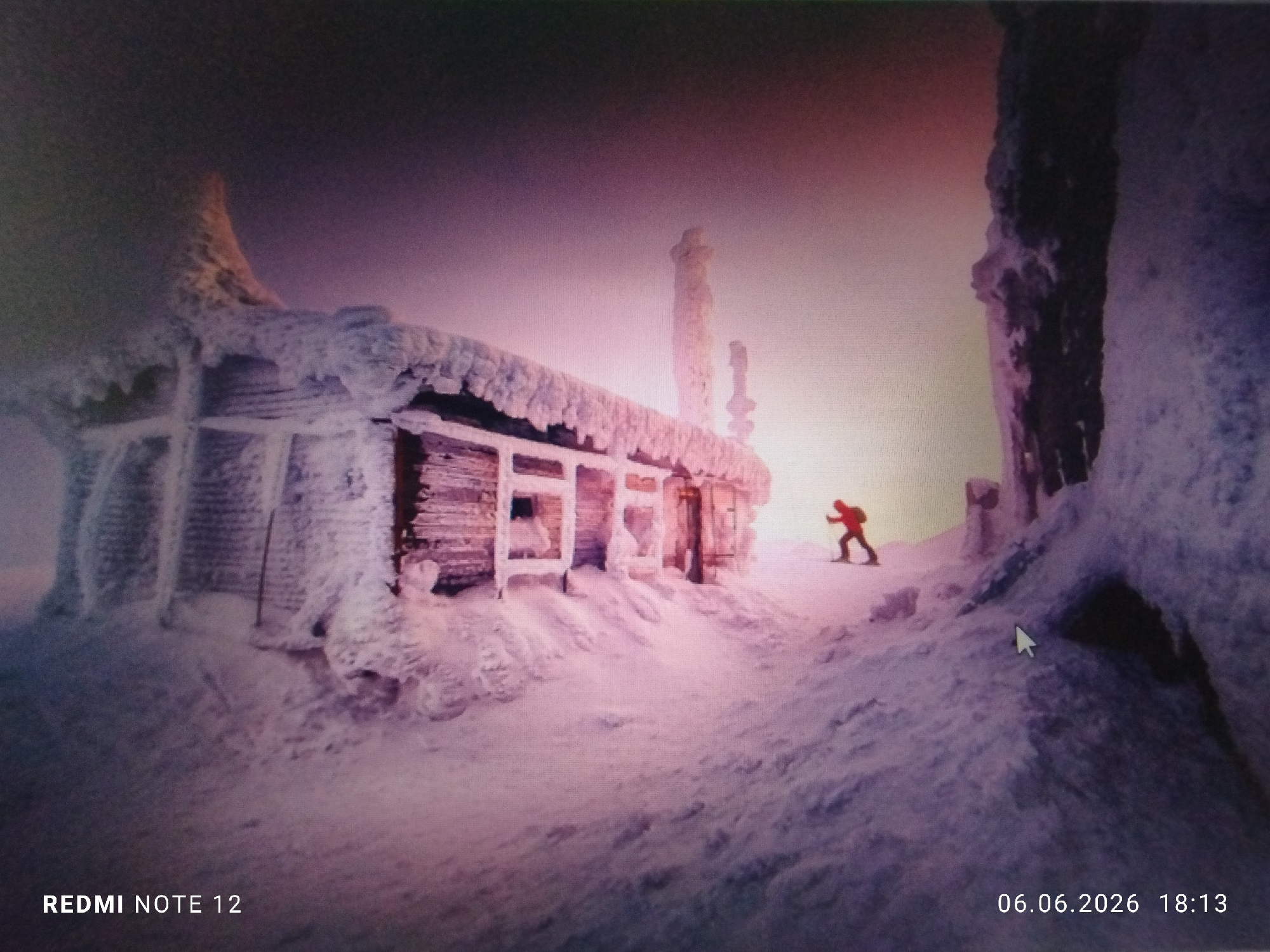The image depicts an extremely cold, snow-covered landscape within the city of Burgas, Bulgaria, captured from a digital display, as indicated by visible pixelation and a white mouse cursor. The environment is dominated by thick layers of rime ice and snow, creating a frigid and desolate atmosphere under a sky transitioning from dark tones in the upper left to a bright, pinkish-orange hue in the lower right, suggesting either dawn or dusk.
The central subject is a single-story, rectangular wooden building, likely a mountain hut or shelter, which is heavily encrusted with white snow and ice, obscuring much of its original structure. The roof, window frames, and walls are caked in thick, sculpted ice, giving the building an organic, almost frosted appearance. Several tall, irregular formations of ice or snow-covered objects, possibly chimneys or natural pinnacles, protrude from the roofline and directly behind the structure.
To the right of the building, a solitary individual in a bright red jacket and dark lower garments is observed moving through deep snow. The person appears to be walking uphill, utilizing two poles, possibly for skiing or trekking, against the stark white backdrop. The ground around the building and where the person walks is entirely covered in undulating snowdrifts and ice, with no bare ground visible.
In the bottom left corner of the image, the text "REDMI NOTE 12" is digitally overlaid. In the bottom right corner, the date and time "06.06.2026 18:13" are displayed, along with a white mouse cursor visible slightly above and to the left of the timestamp.