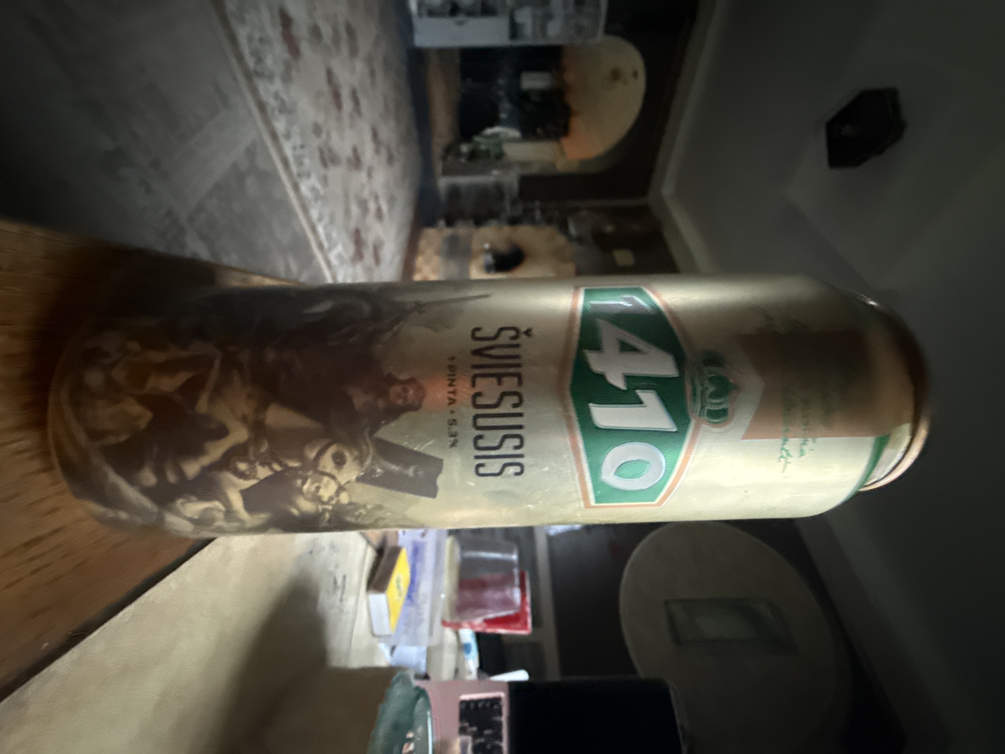 The photograph shows a can of beer labeled "ŠVIESUSIS 1410" positioned on a wooden table. The can has a predominantly gold and green design with a graphic depicting a historical scene. In the background, out of focus, are elements of an interior space, including a carpeted floor, what appears to be an archway, and possibly a fireplace or oven structure. The lighting is dim, and the overall impression is of an indoor setting, likely a room in a residence or a tavern. No specific location in the city of Galinė, Lithuania, can be identified from the visual information provided. The image is a still photograph with no people or animals actively engaged in any interaction.