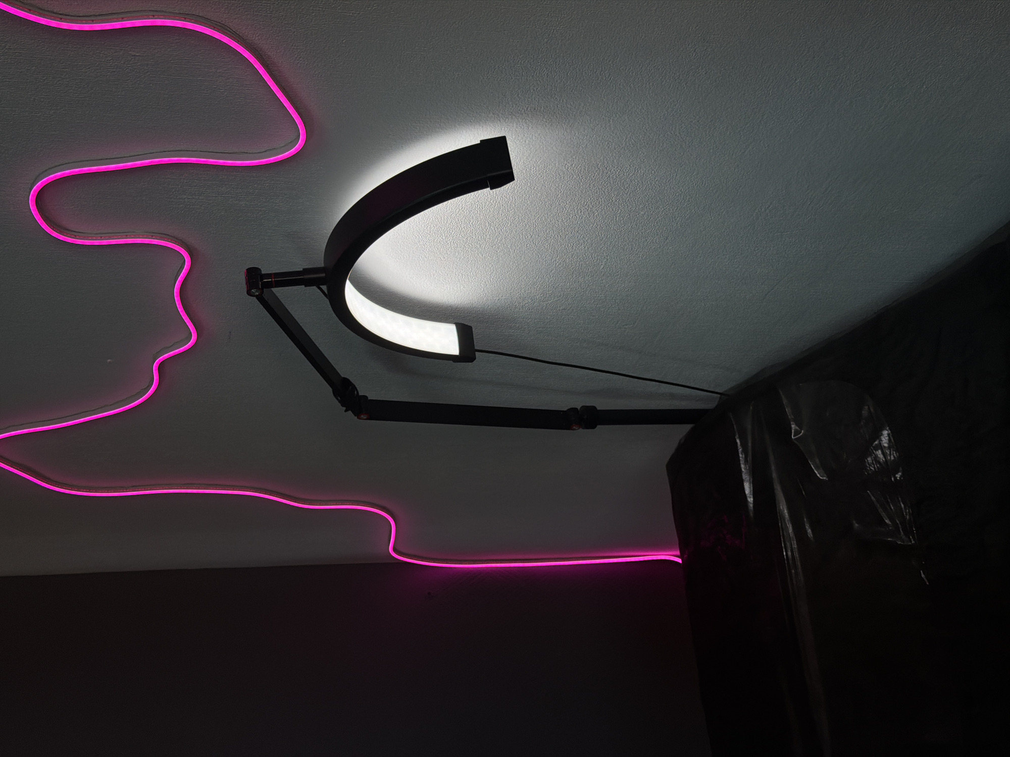 The image shows an interior view of a room, likely in a private residence, with a white ceiling and dark walls. A hot pink neon light fixture runs along the ceiling, creating a wavy pattern. A black adjustable arm lamp with a curved LED light is mounted to the ceiling. The lamp is illuminated, casting light on the ceiling. A dark, opaque object, possibly covered by a plastic sheet, is positioned in the lower right corner. The location in the city of Chaikovsky, Russia is not readily discernible from the image alone.