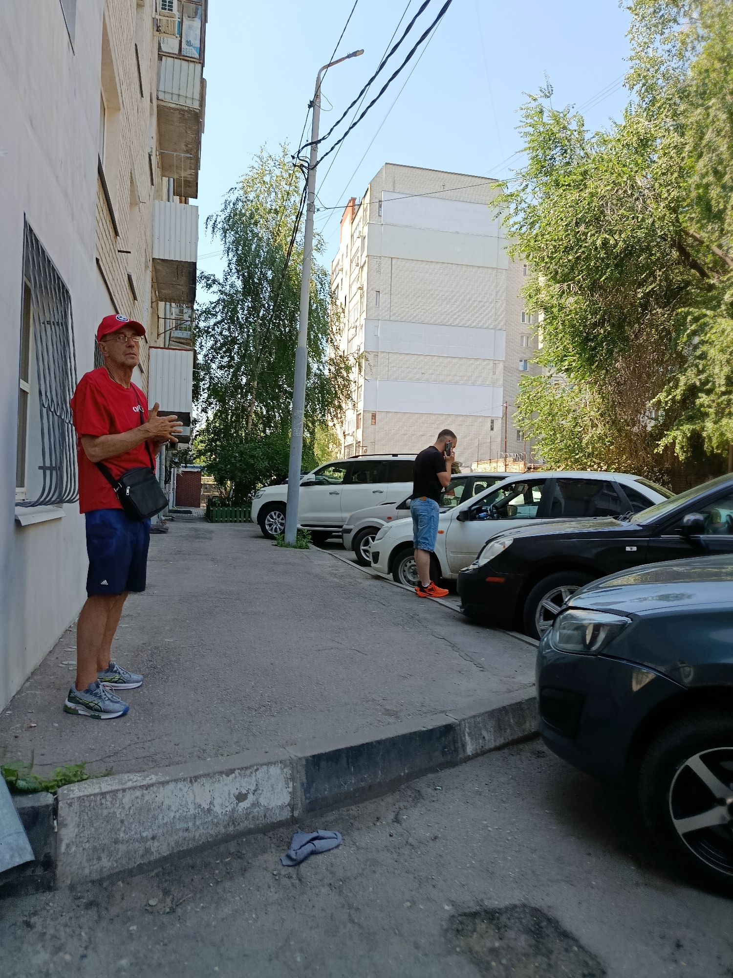 A man in a red shirt and blue shorts stands on a sidewalk next to a white building. He is wearing glasses and a red baseball cap, and he has a black bag slung across his chest. His hands are clasped in front of him. Another man in a black shirt and blue shorts is standing by several parked cars, looking at his phone. The scene appears to be in a city, with buildings and trees visible in the background. There are multiple cars parked along the street. The lighting suggests it is daytime.