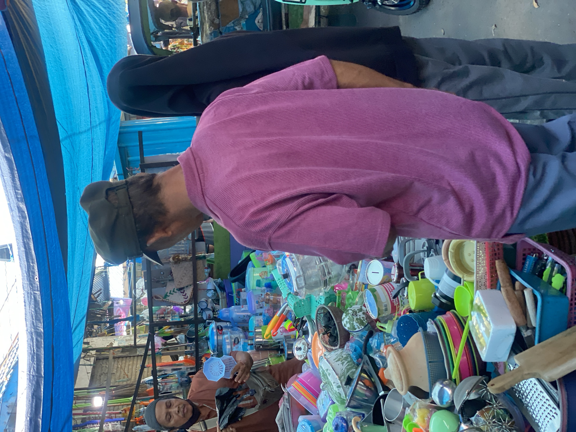 This image depicts a scene at an outdoor market in Desa Tunggul Irang, Indonesia. In the foreground, a man wearing a pink collared shirt and a green cap is facing away from the camera, appearing to look at items on display. To his left, a woman wearing a brown headscarf and a brown shirt is holding up a black plastic bag and a blue basket. She is also wearing a dark face mask. Behind the man in the pink shirt, another person is visible, almost entirely covered in black, with only a small portion of their face showing.
The market stall is laden with a variety of household items, primarily plastic containers and kitchenware. There are numerous colorful plastic bowls, plates, cups, and storage containers in shades of pink, blue, green, and yellow. Metal pots and pans are also visible, along with a wooden cutting board and a grater. A blue tarp is stretched overhead, providing shade. The background shows more market stalls with various goods, including what appear to be brooms and cleaning supplies. The ground is a paved surface, possibly asphalt, with some fallen leaves. The overall impression is a busy, everyday marketplace.